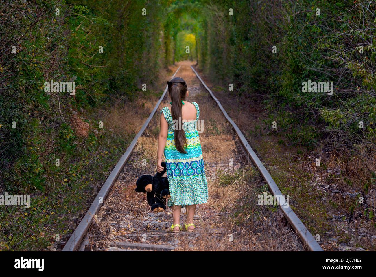 Petite fille à l'arrière, vêtue d'une longue robe tenant un ours en peluche debout sur les vieux rails dans un labyrinthe de tunnel ferroviaire abandonné. Banque D'Images