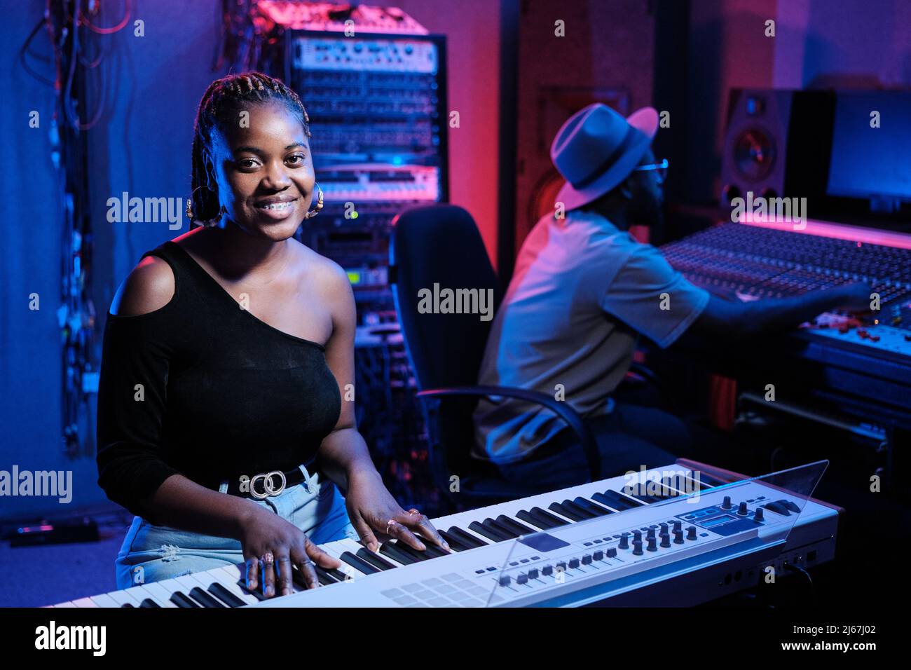 Joyeuse jeune musicienne afro-américaine jouant du clavier numérique tout en travaillant sur la console de mixage dans un studio d'enregistrement Banque D'Images