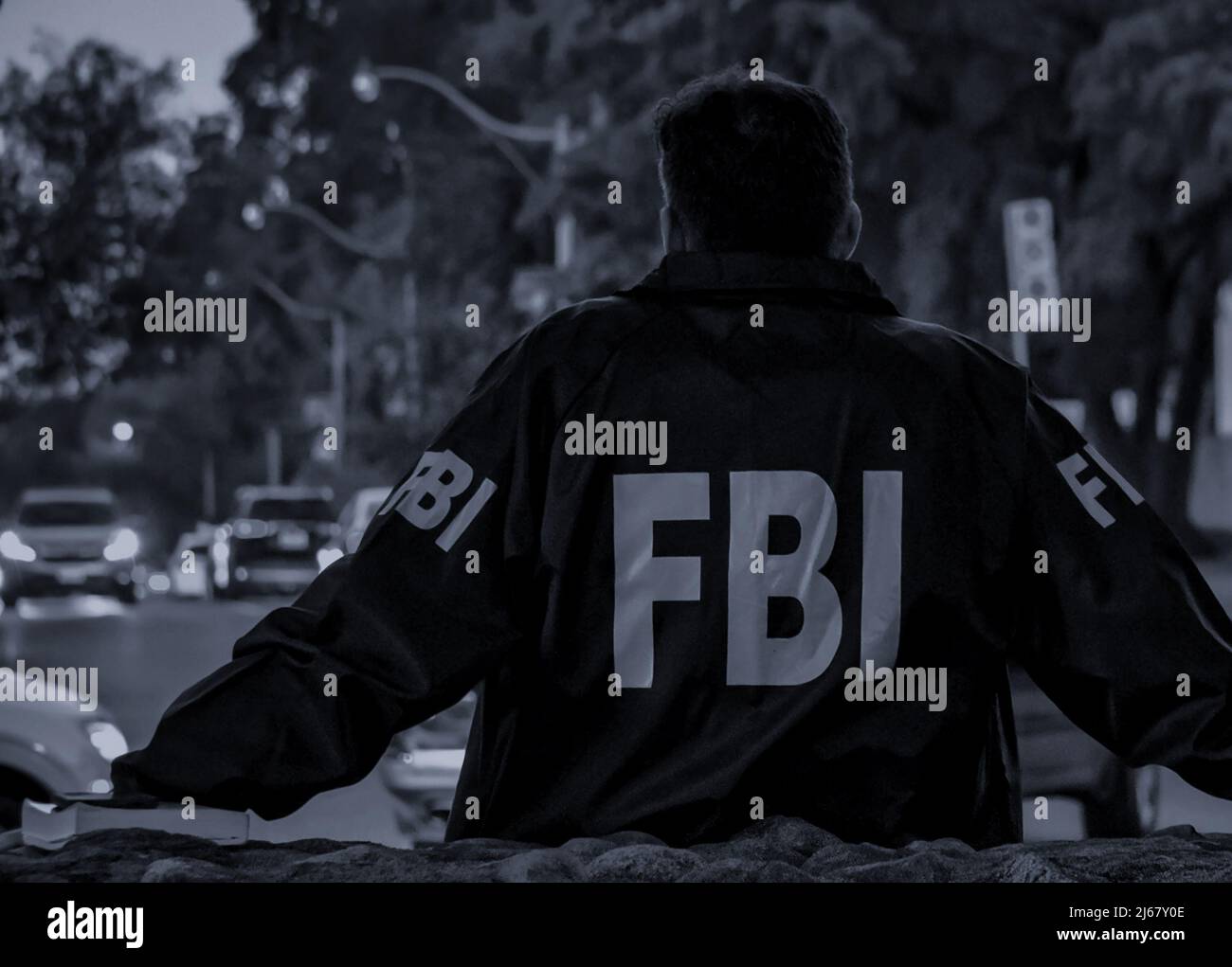 L'agent FBI mâle porte un manteau bleu foncé avec le logo FBI regardant dans la rue avec des voitures au crépuscule vu de derrière. Banque D'Images