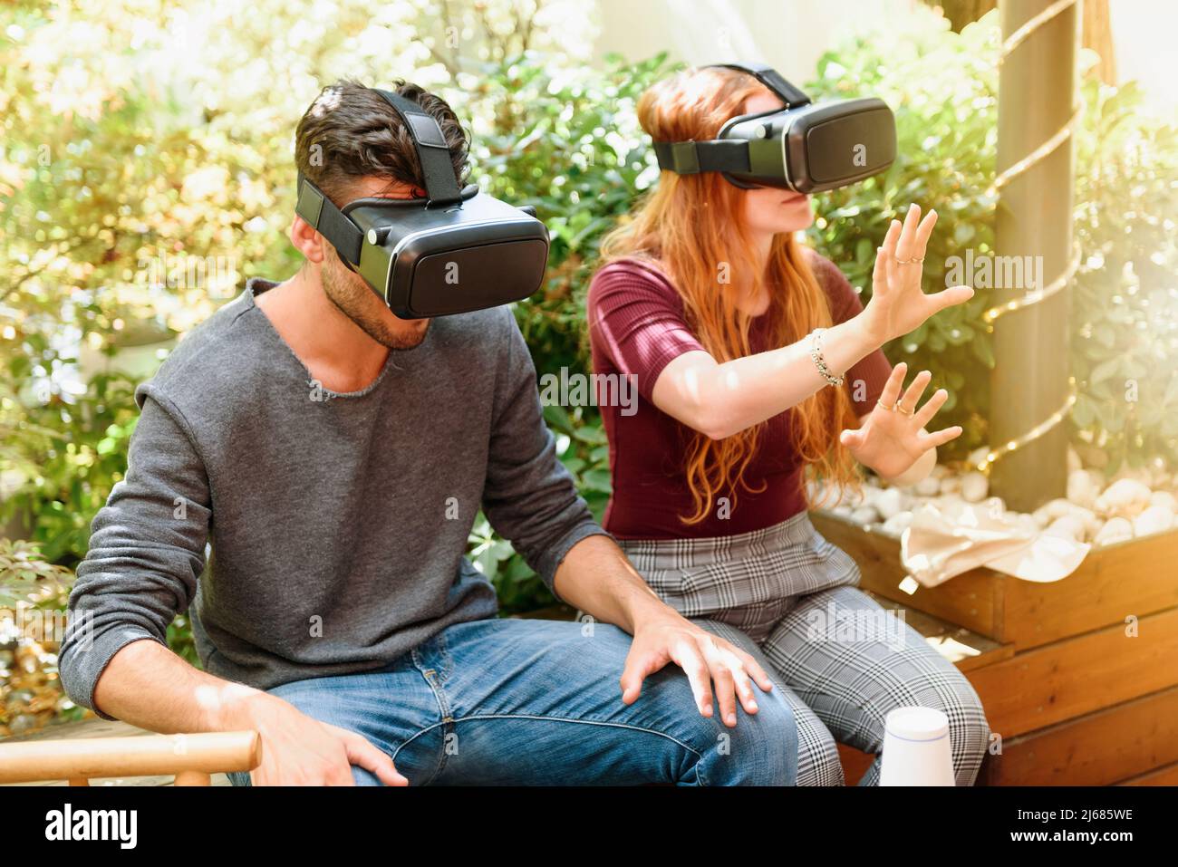 Un couple concentré en vêtements décontractés assis sur un banc en bois sur la terrasse et utilisant des visières VR contre des buissons verts par temps ensoleillé Banque D'Images