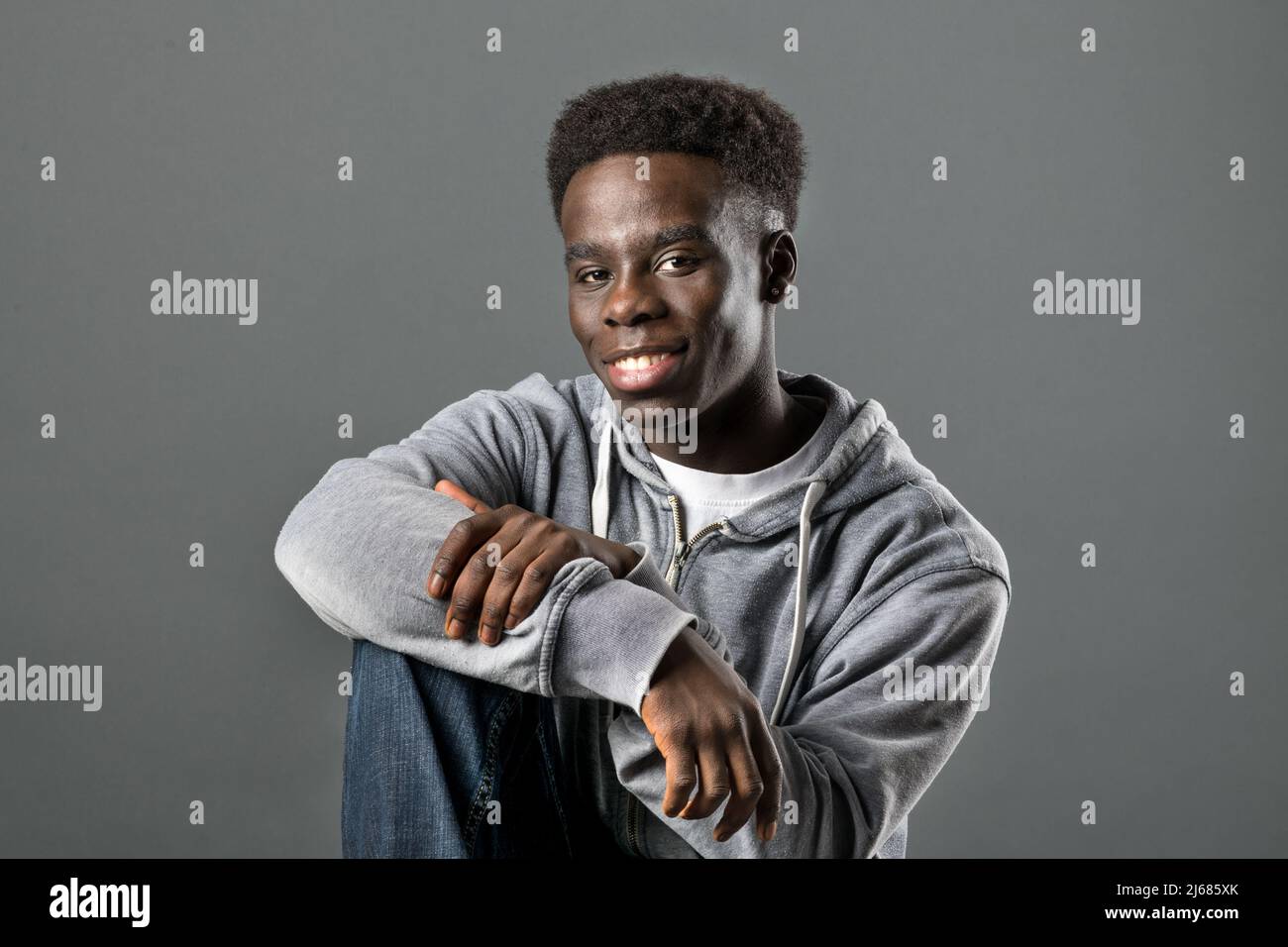 Positif Afro-américain homme portant un pull à capuche décontracté et un Jean assis en studio sur fond gris et regardant l'appareil photo Banque D'Images