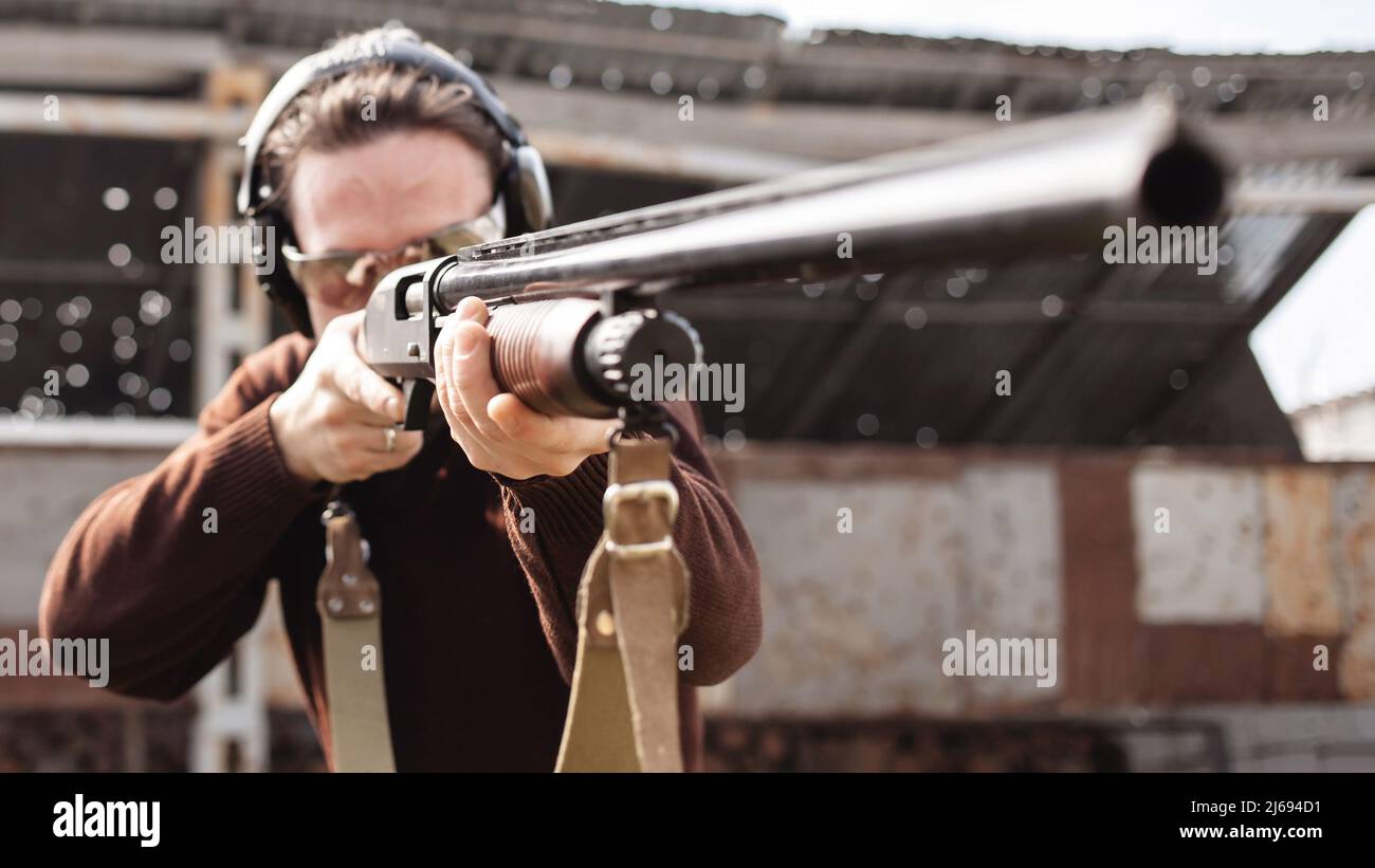 Un jeune homme dans des lunettes de protection et des écouteurs. Un fusil à pompe. Pneus à l'extérieur. Un mur et un toit avec trous de balle. Sport Banque D'Images