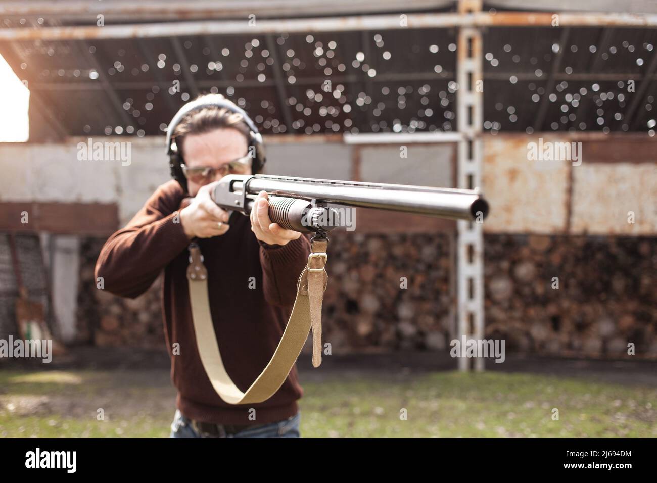 Un jeune homme dans des lunettes de protection et des écouteurs. Un fusil à pompe. Pneus à l'extérieur. Un mur et un toit avec trous de balle. Sport Banque D'Images