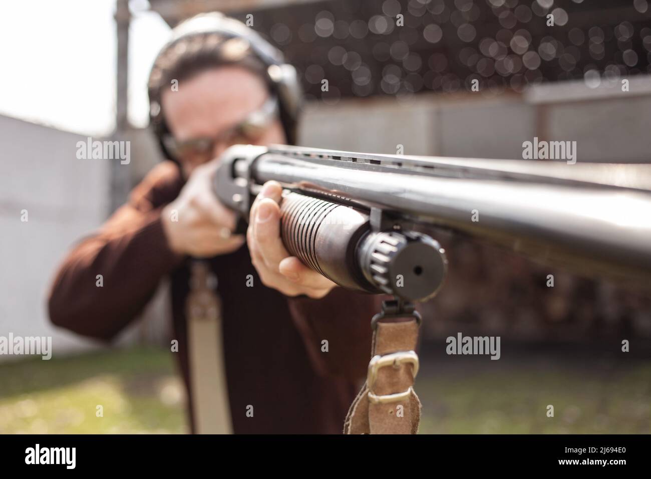 Un jeune homme dans des lunettes de protection et des écouteurs. Un fusil à pompe. Pneus à l'extérieur. Un mur et un toit avec trous de balle. Sport Banque D'Images