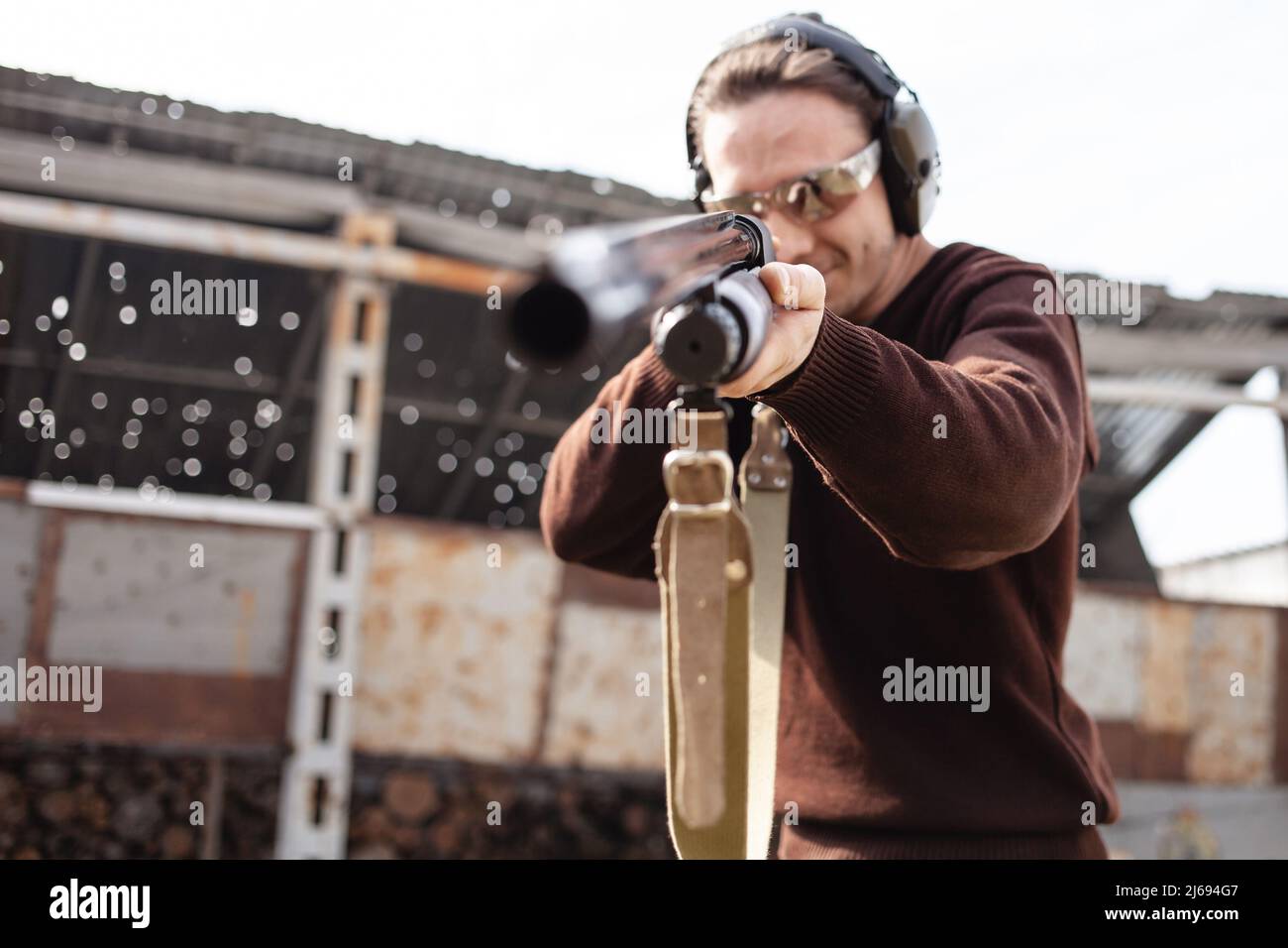 Un jeune homme dans des lunettes de protection et des écouteurs. Un fusil à pompe. Pneus à l'extérieur. Un mur et un toit avec trous de balle. Sport Banque D'Images