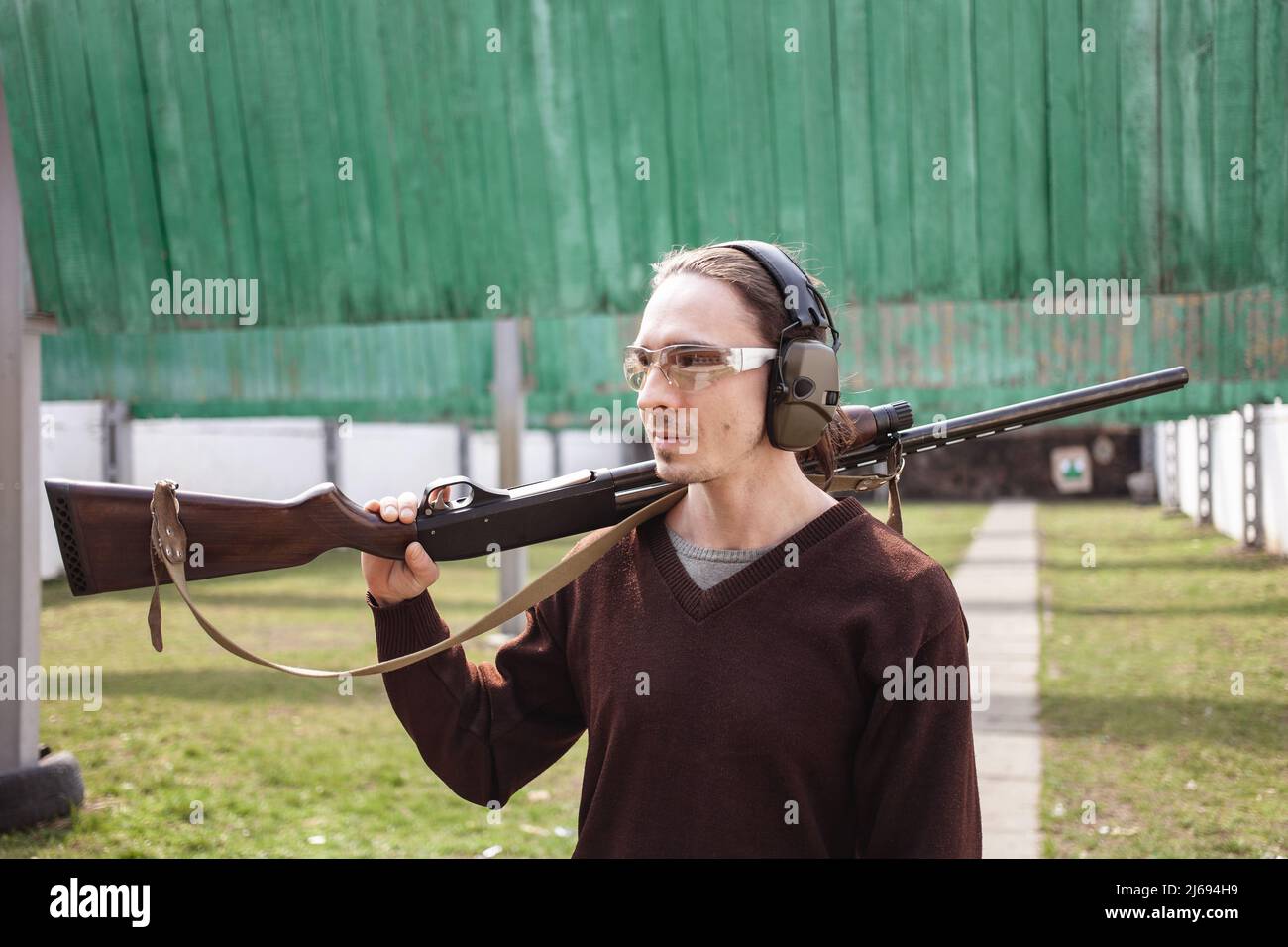 Un jeune homme dans des lunettes de protection et des écouteurs. Un fusil à pompe. Pneus à l'extérieur. Herbe verte, protection pour le tournage de sports. Hobby. Banque D'Images