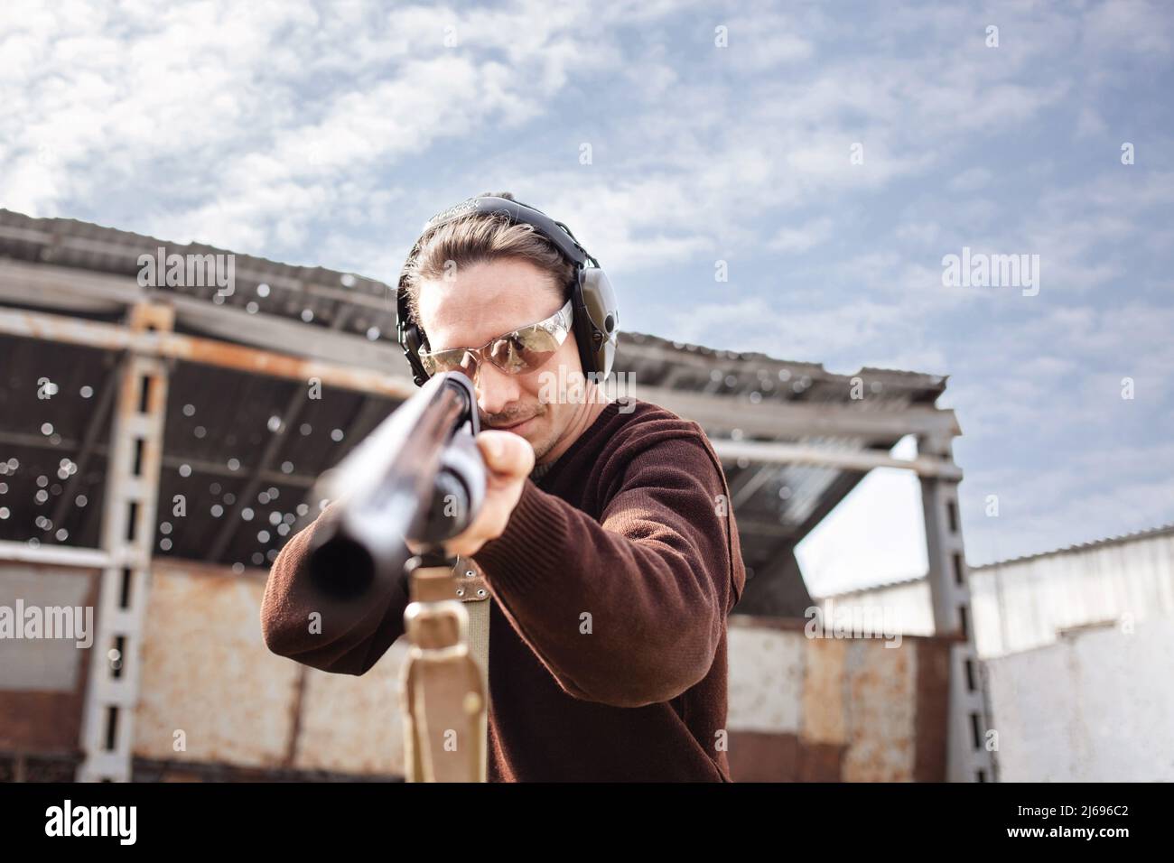 Un jeune homme dans des lunettes de protection et des écouteurs. Un fusil à pompe. Pneus à l'extérieur. Un mur et un toit avec trous de balle. Sport gros plan Banque D'Images