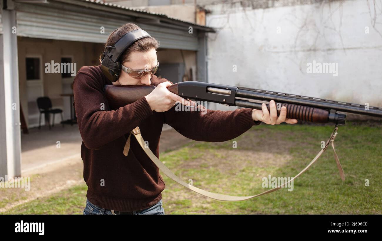 Un jeune homme avec une arme à feu pompeuse sur fond blanc. Un homme dans des lunettes de protection et des écouteurs. Pneu extérieur. Pistolet hangar gris Banque D'Images