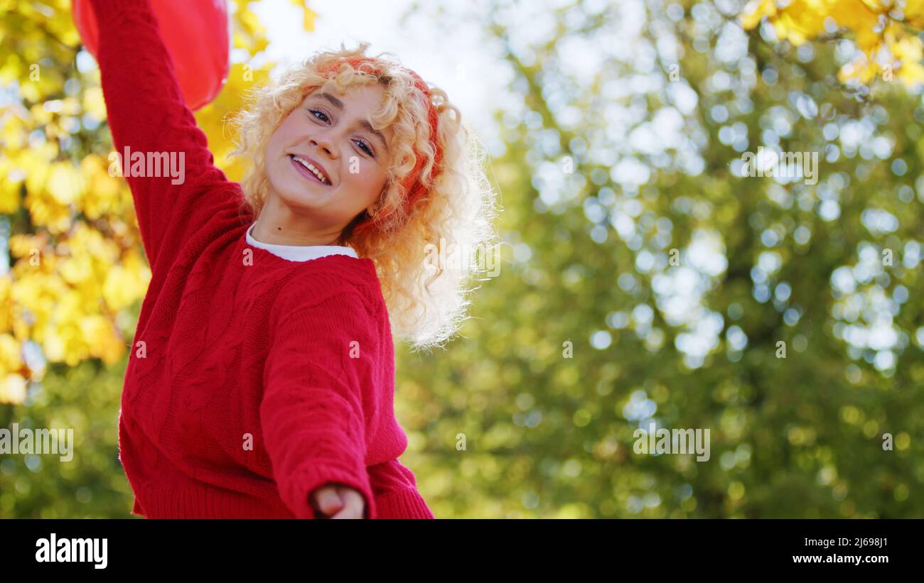 Jolie petite fille caucasienne mignonne avec de beaux cheveux blonds dans un chandail rouge regardant la caméra, dansant, tenant le ballon rouge. Prise de vue en extérieur. Photo de haute qualité Banque D'Images