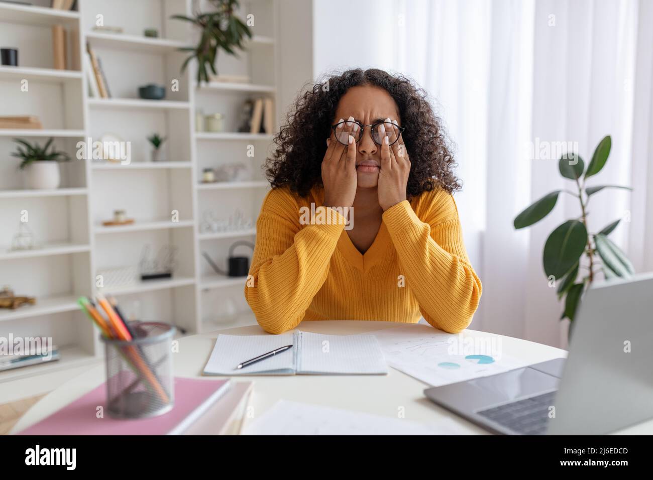 Jeune femme noire en lunettes frottant les yeux irrités, assis au bureau avec un ordinateur portable, étudiant trop en ligne à la maison Banque D'Images