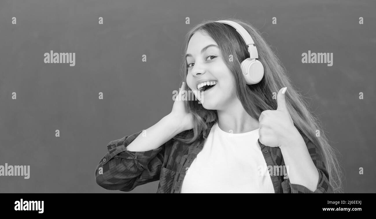 bonne fille tient des écouteurs modernes à l'école secondaire au tableau noir montrant le pouce vers le haut, accessoire audio. Banque D'Images