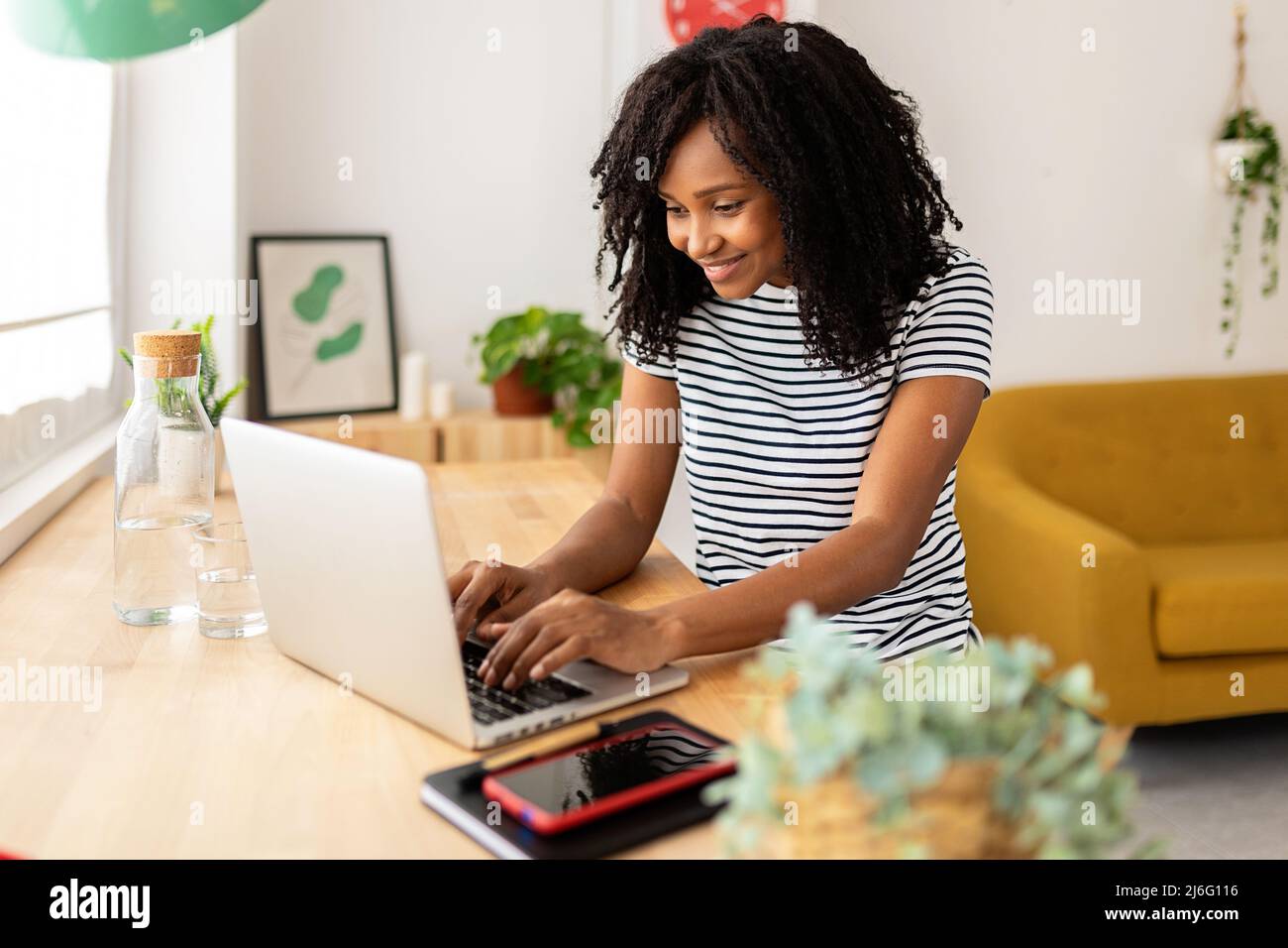 Jeune femme travaillant sur son ordinateur portable tout en étant assise sur un bureau à la maison. Banque D'Images
