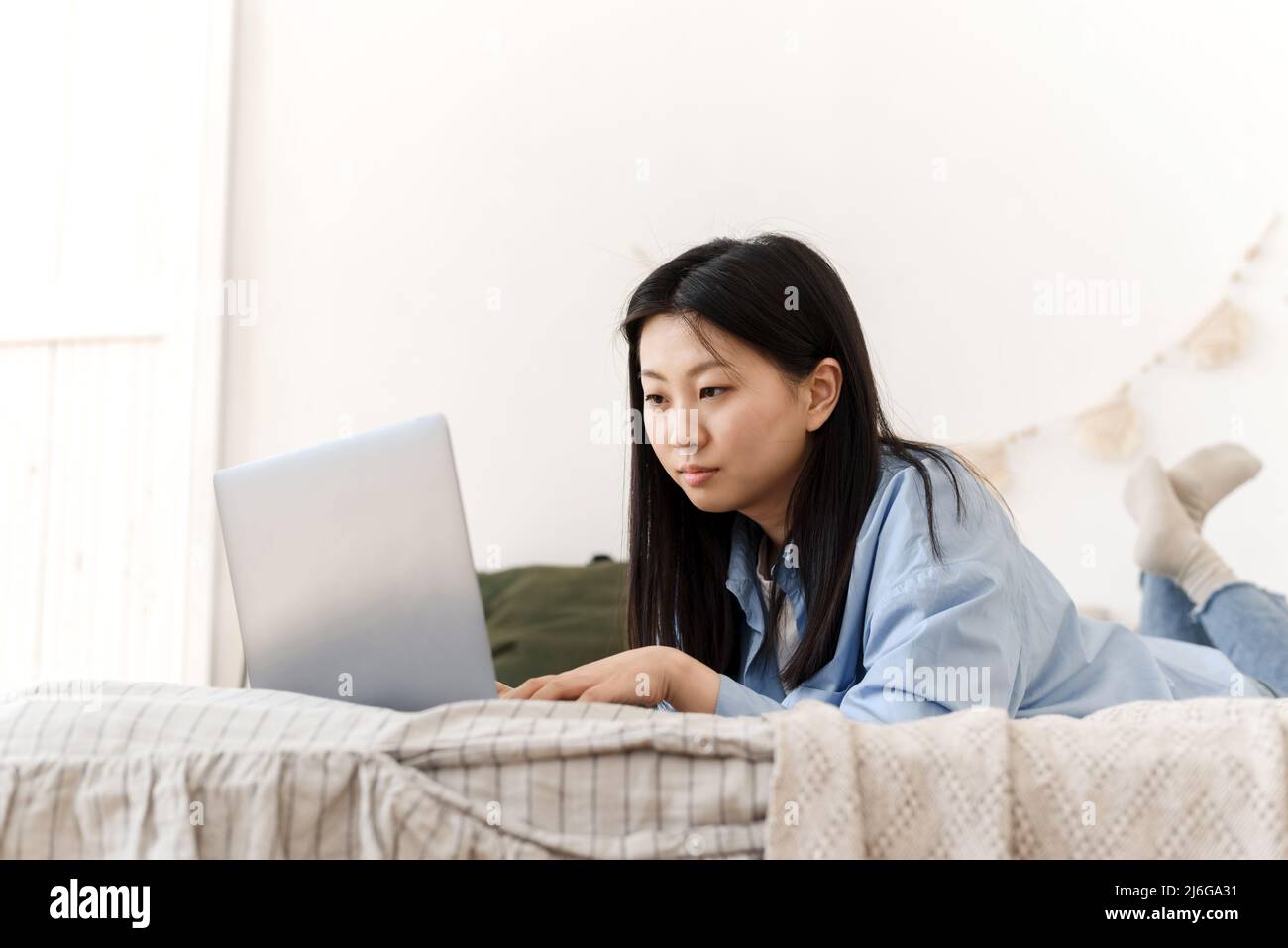 Une belle femme asiatique est allongée sur le lit et travaille à l'aide d'un ordinateur portable de la maison. Une jeune femme travaille à la maison en quarantaine Banque D'Images