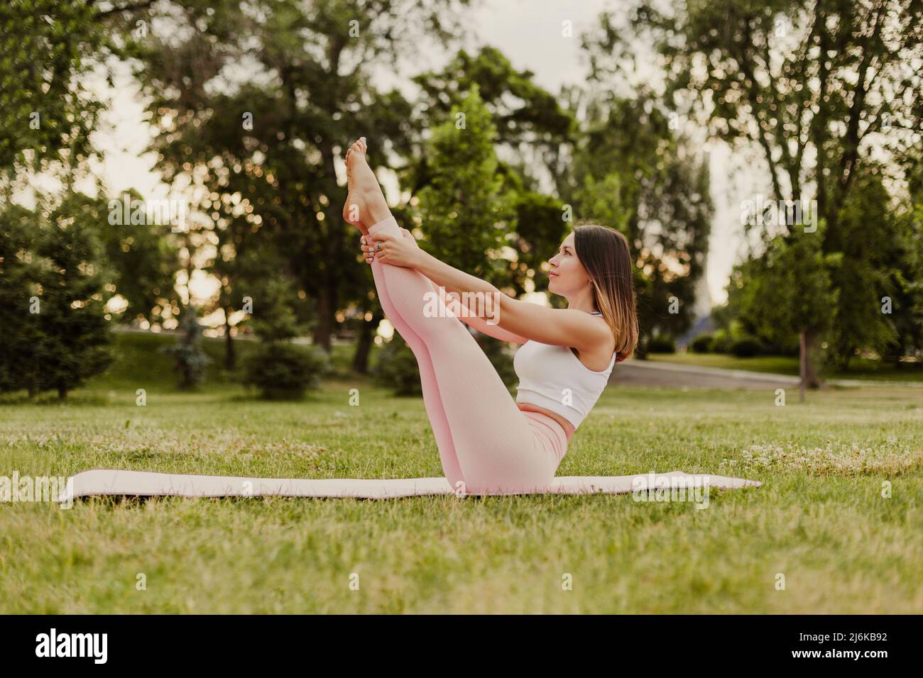 Jeune femme en forme pratiquer le yoga sur tapis de gymnastique dans le parc vert sur l'herbe. Tenez-vous debout dans la posture du bateau dans les vêtements de sport, faites des exercices pour garder la forme le matin de l'été Banque D'Images