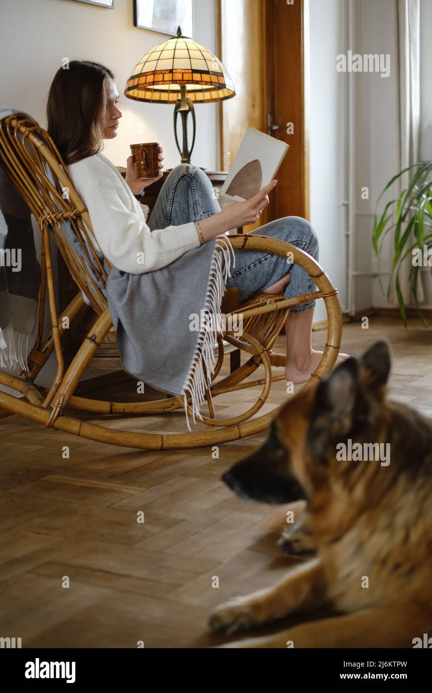 Jeune femme lisant un livre avec chien Berger allemand, assise sur une chaise à bascule. Ambiance chaleureuse, vie lente, personnes avec animaux de compagnie. Maison d'époque Banque D'Images