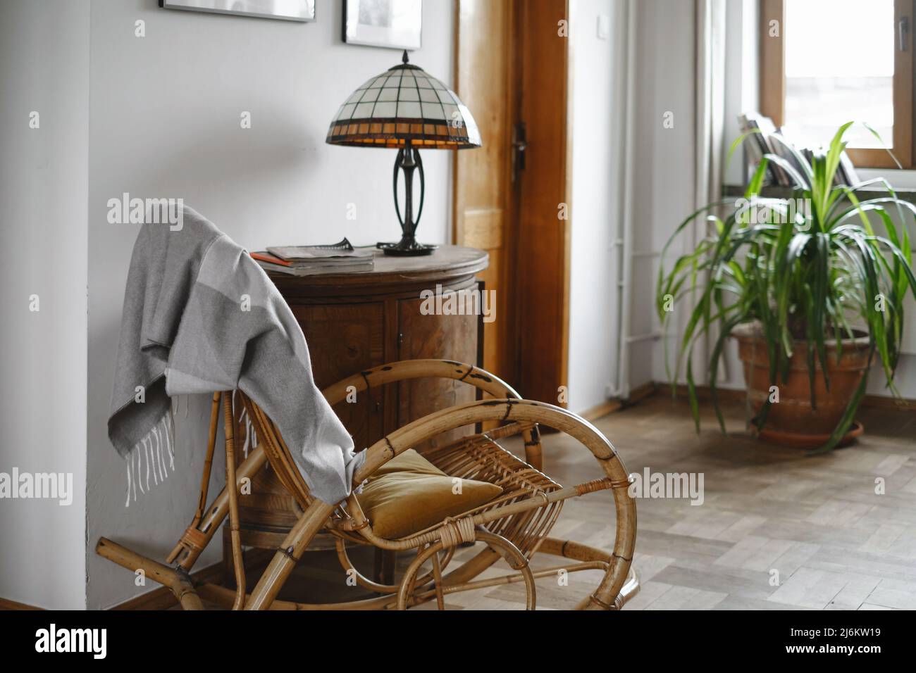 Fauteuil à bascule en osier de style ancien avec couverture dans une maison confortable, lampe sur armoire en bois. Lieu de lecture, pas de personnes, usine à la maison près de la fenêtre. Confortable Banque D'Images