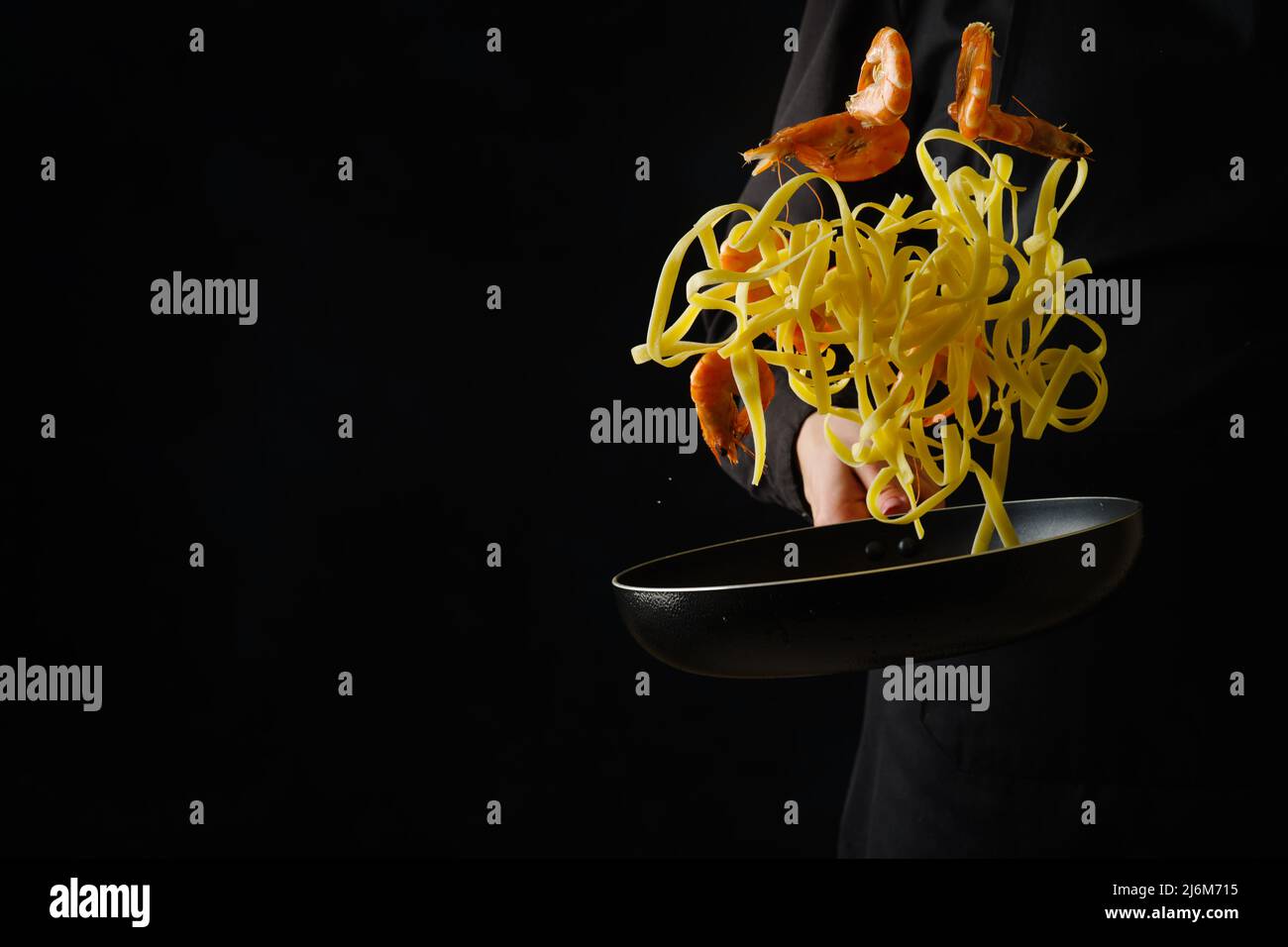 Cuisine italienne. Pâtes de crevettes dans une poêle sur fond noir dans un vol gelé dans la main d'un chef professionnel en uniforme noir. Publicité Banque D'Images