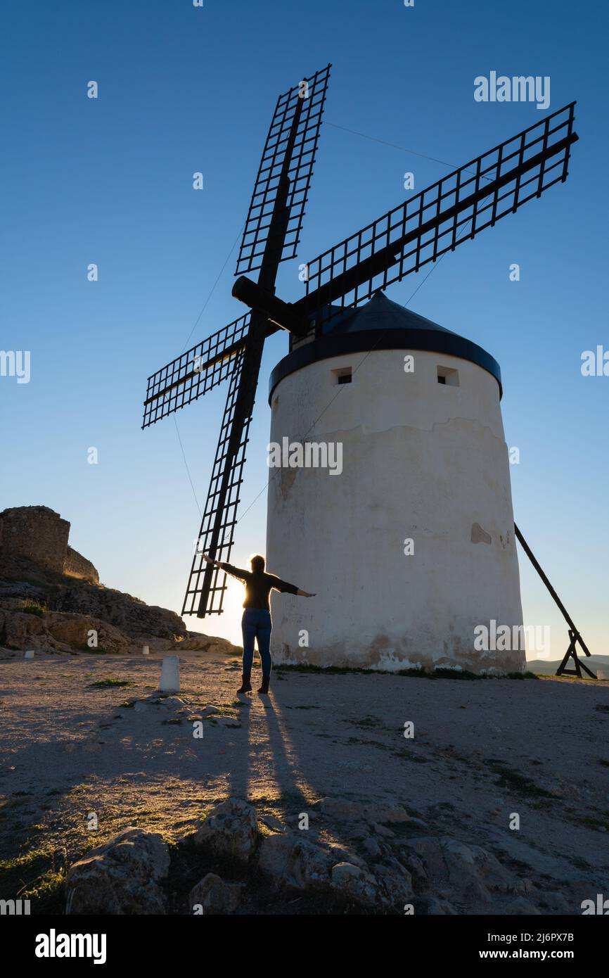 Moulin à vent blanc avec ses lames en bois, au coucher du soleil dans une campagne castillane en Espagne et un ciel bleu, avec une femme levant les mains imitant la blad Banque D'Images