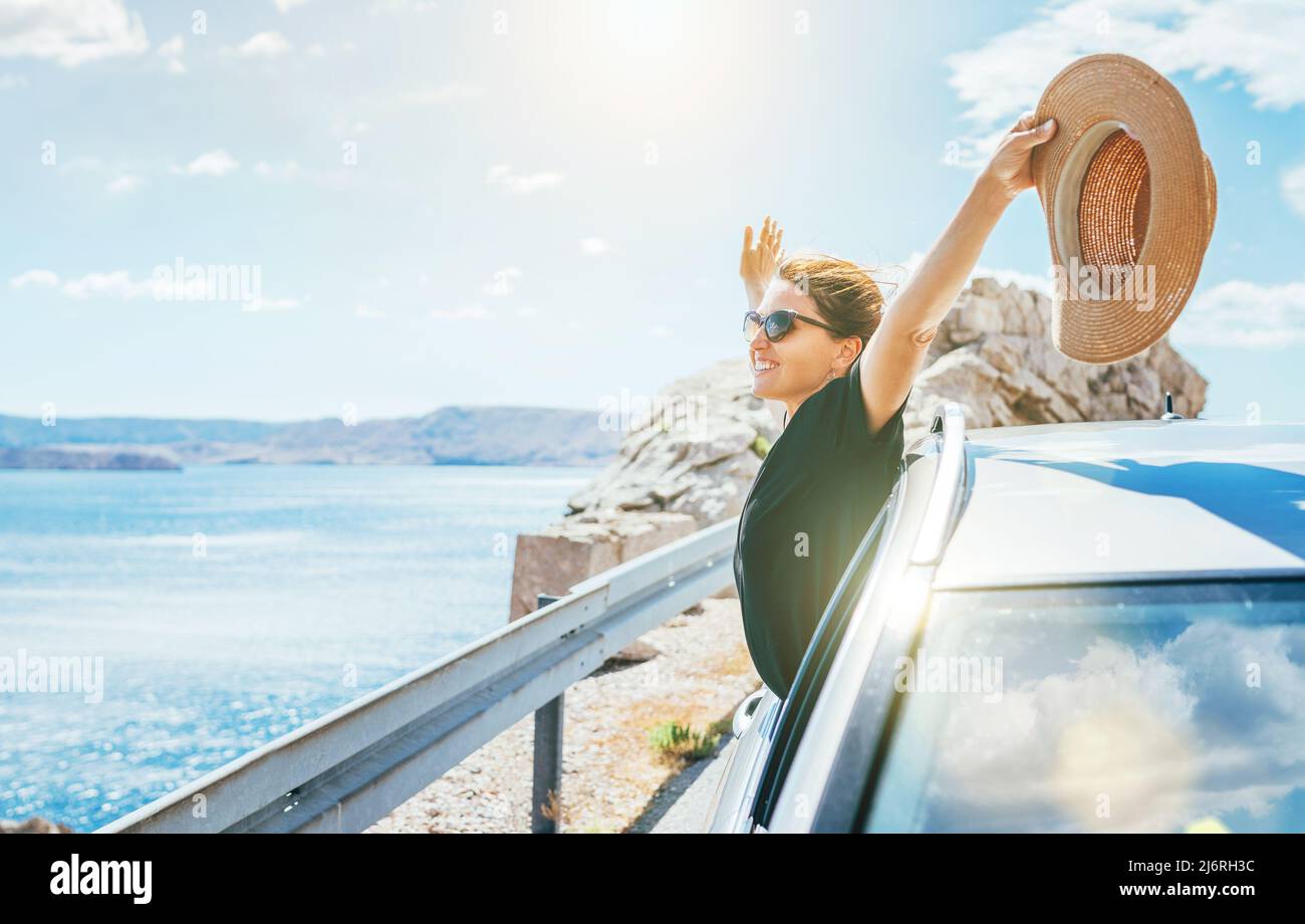 Portrait de femme joyeuse appréciant le voyage en bord de mer. Habillée d'une robe noire avec chapeau de paille et lunettes de soleil elle a ouvert les bras et brillant avec happ Banque D'Images