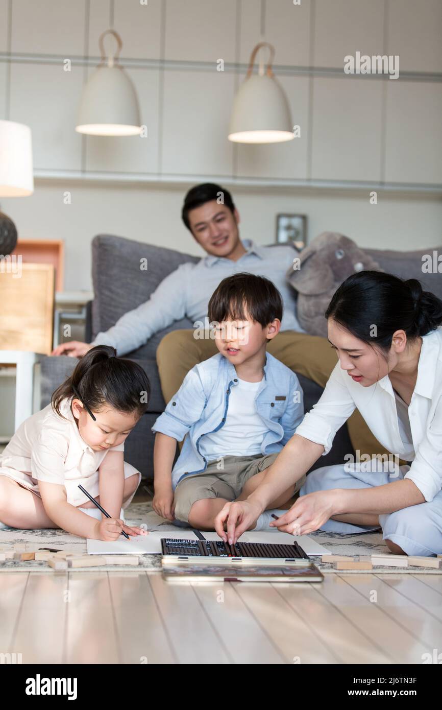 Une jeune mère assise patiemment sur un tapis avec des enfants dessinant des photos au crayon, tandis que le père est confortablement assis sur un canapé - photo de stock Banque D'Images