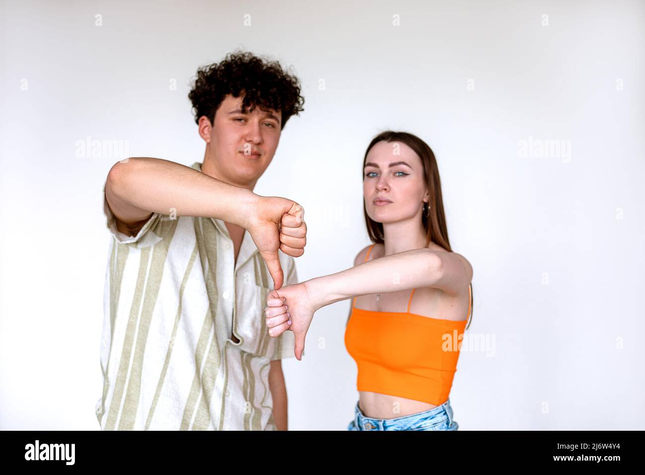 Portrait d'un jeune couple sérieux posant sur fond blanc. Jeune homme bouclés confiant, femme montrant le signe du pouce vers le bas. Banque D'Images