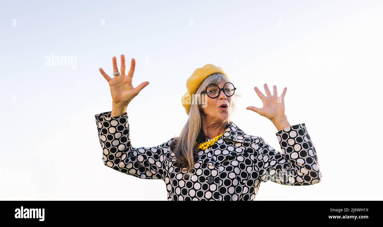 Femme âgée soulevant ses mains en jeu tout en se tenant debout à l'extérieur. Femme âgée blissful portant des vêtements décontractés colorés en été. Tapis insouciant Banque D'Images