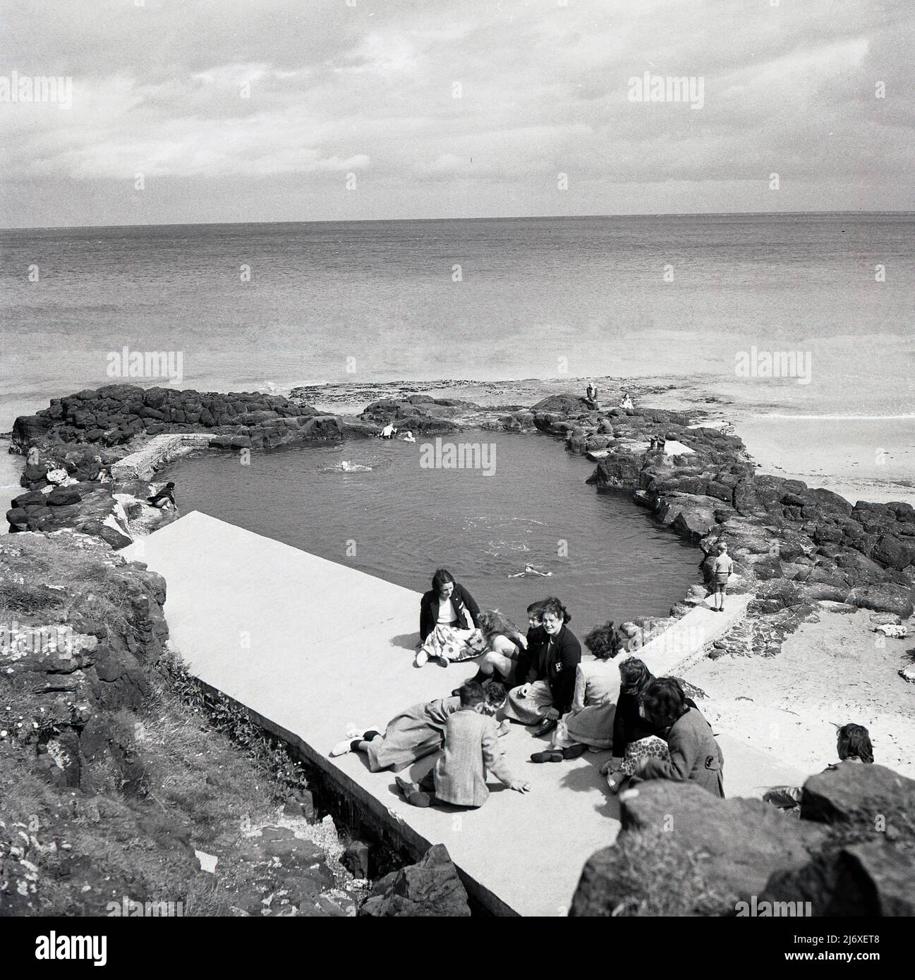 1950s, historique, une mère et des enfants d'école sérale assis sur une corniche de béton près d'une piscine côtière naturelle sur la côte d'Antrim, Irlande du Nord, Royaume-Uni. Banque D'Images