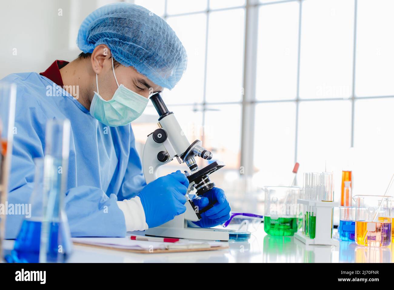Un chercheur asiatique en laboratoire ou en pharmacie note tout en étudiant un échantillon de virus au microscope Banque D'Images