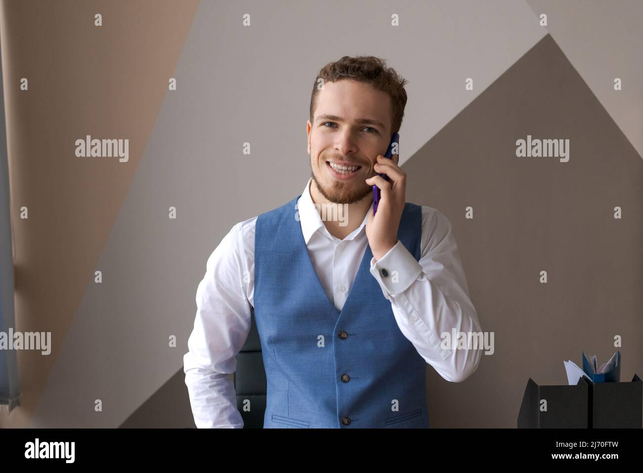 Agréable beau barbu homme en chemise et gilet parler au téléphone, commander un taxi ou la livraison de nourriture au bureau au travail. Un jeune homme d'affaires caucasien souriant tient un appel avec des clients partenaires. Banque D'Images