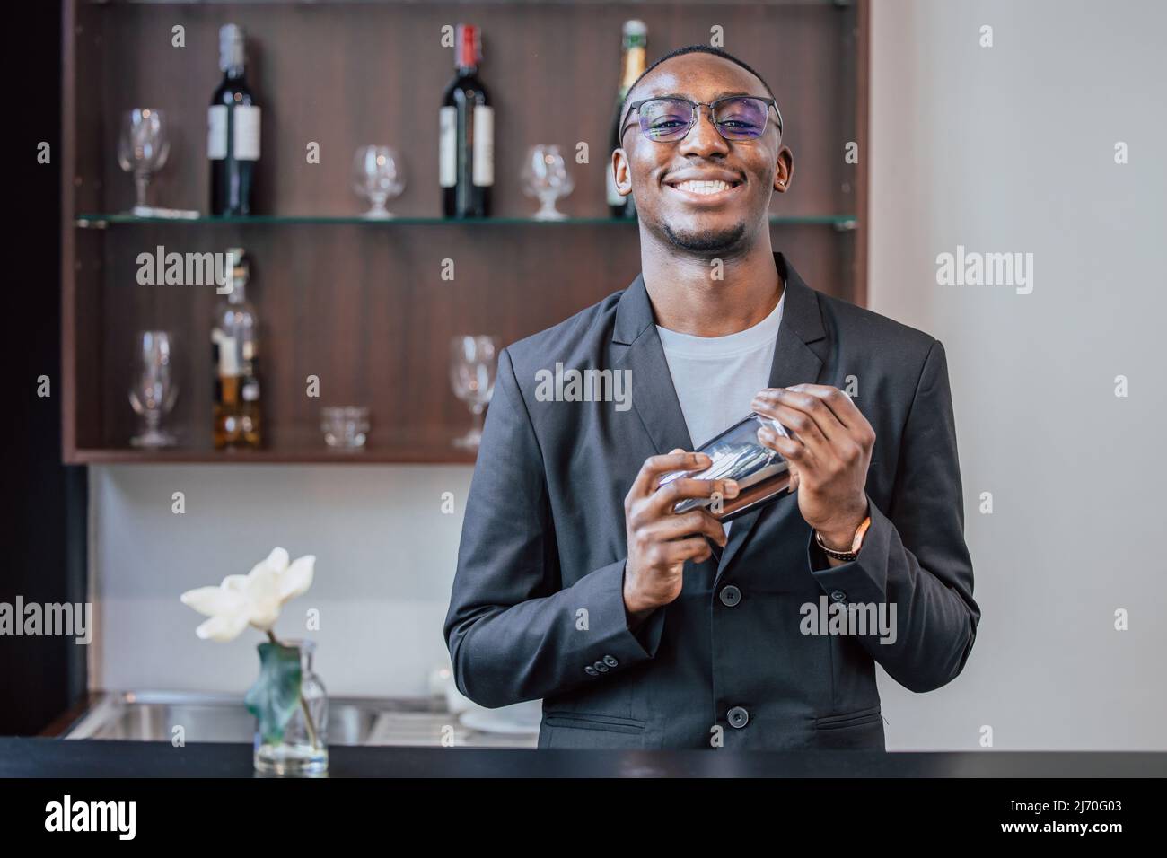 un jeune barman souriant dans des gens noirs travaille avec un shaker à cocktail dans le luxueux bar-salon du hall de l'hôtel. Banque D'Images