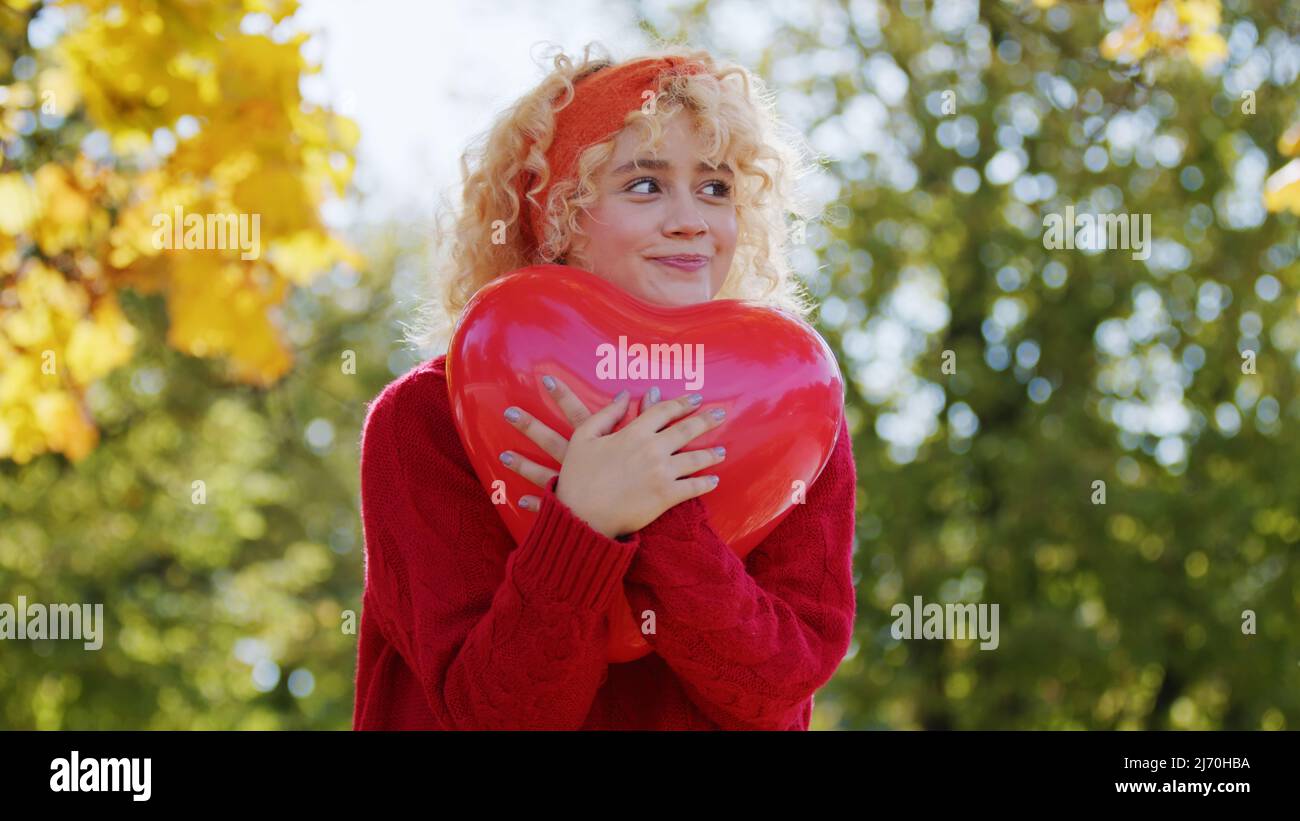 Blonde souriante caucasienne fille tient rouge ballon sous la forme de coeur regardant loin moyenne prise de vue espace de copie arrière-plan du parc . Photo de haute qualité Banque D'Images