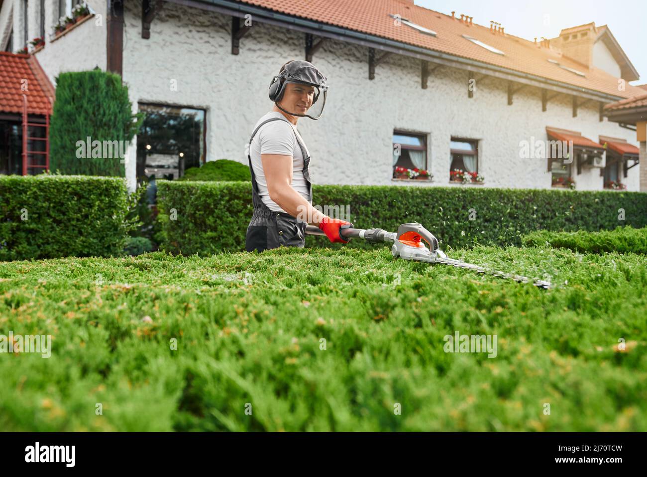 Homme caucasien en uniforme, lunettes de protection et masque de taille des bagues surcultivées avec coupe électrique. Jardinier compétent prenant soin des plantes sur l'arrière-cour pendant l'été. Banque D'Images