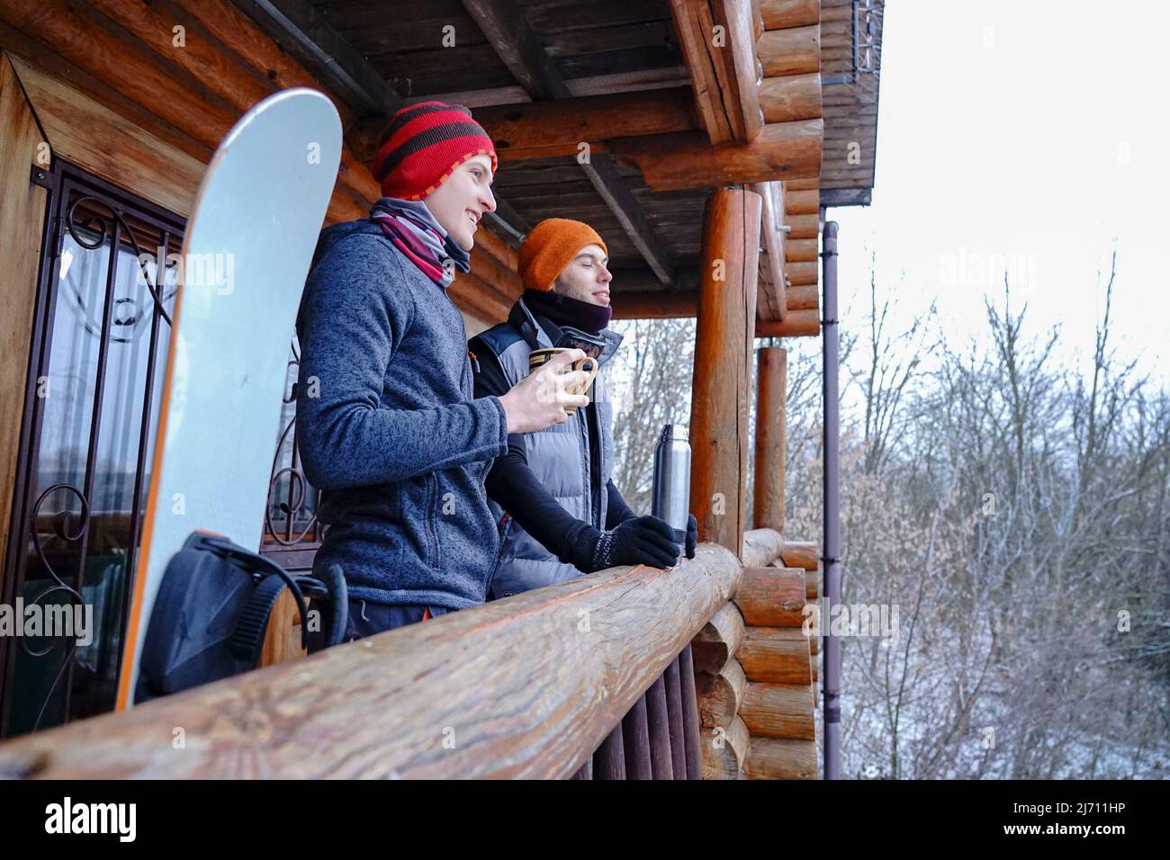 Un gars dans un chapeau d'orange en hiver boit le thé d'un thermos pendant des vacances d'hiver dans les montagnes. Il se tient sur le balcon d'une maison en bois. Port Banque D'Images