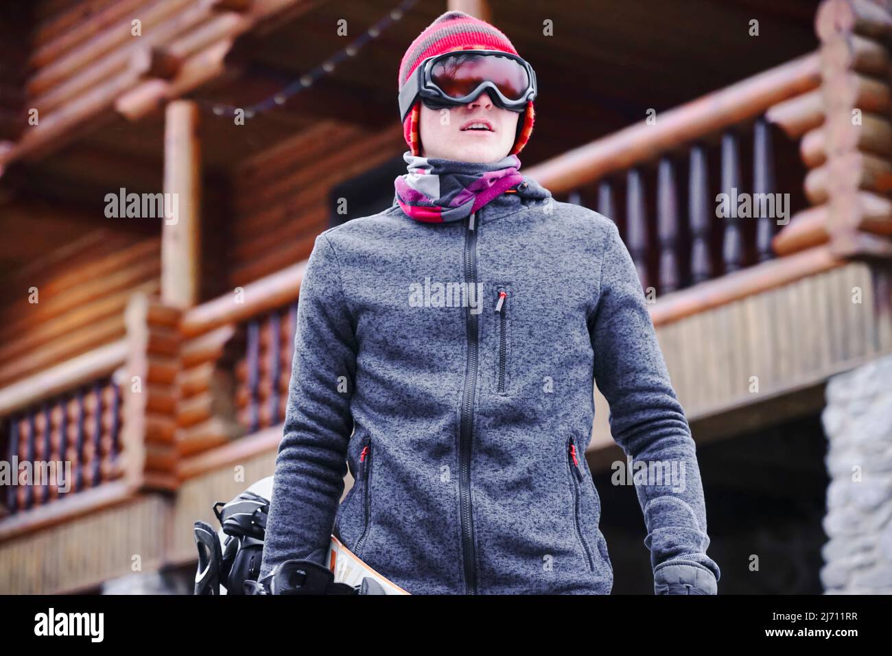Un gars en lunettes de ski tient un snowboard dans la main près de la maison de la station dans la base de ski. Portrait Banque D'Images