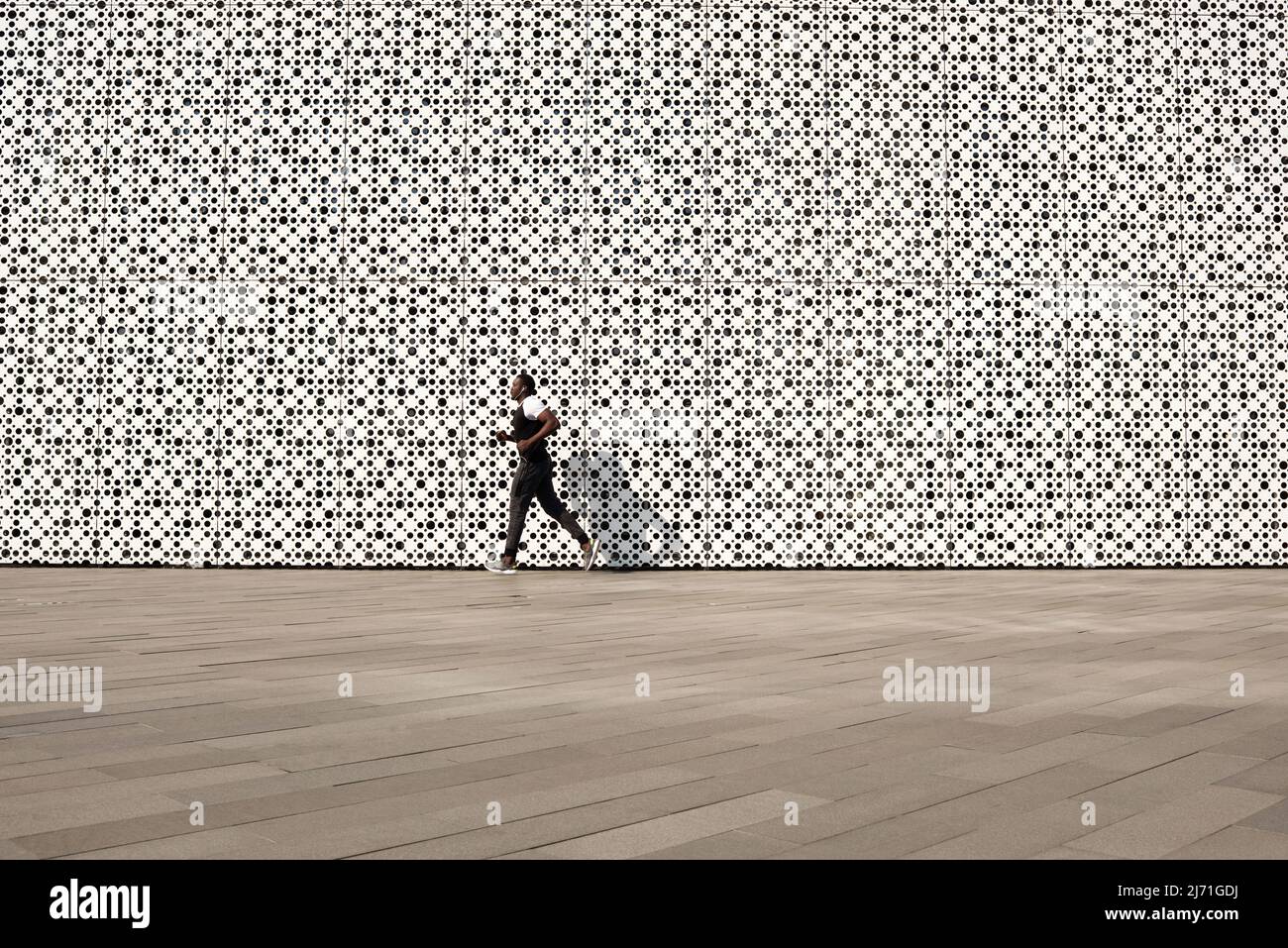 Entraînement en milieu urbain. Sportif noir en forme d'activewear courant le long d'un bâtiment contemporain avec façade perforée blanche, espace copie Banque D'Images