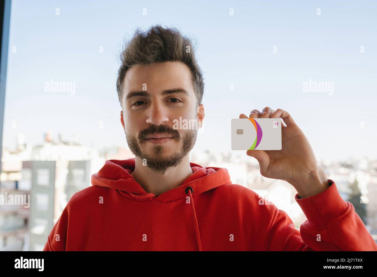 Jeune homme beau portant un sweat à capuche rouge montrant une carte de crédit ou de débit. Achat en ligne ou banque numérique. Accent sélectif sur l'homme. Banque D'Images