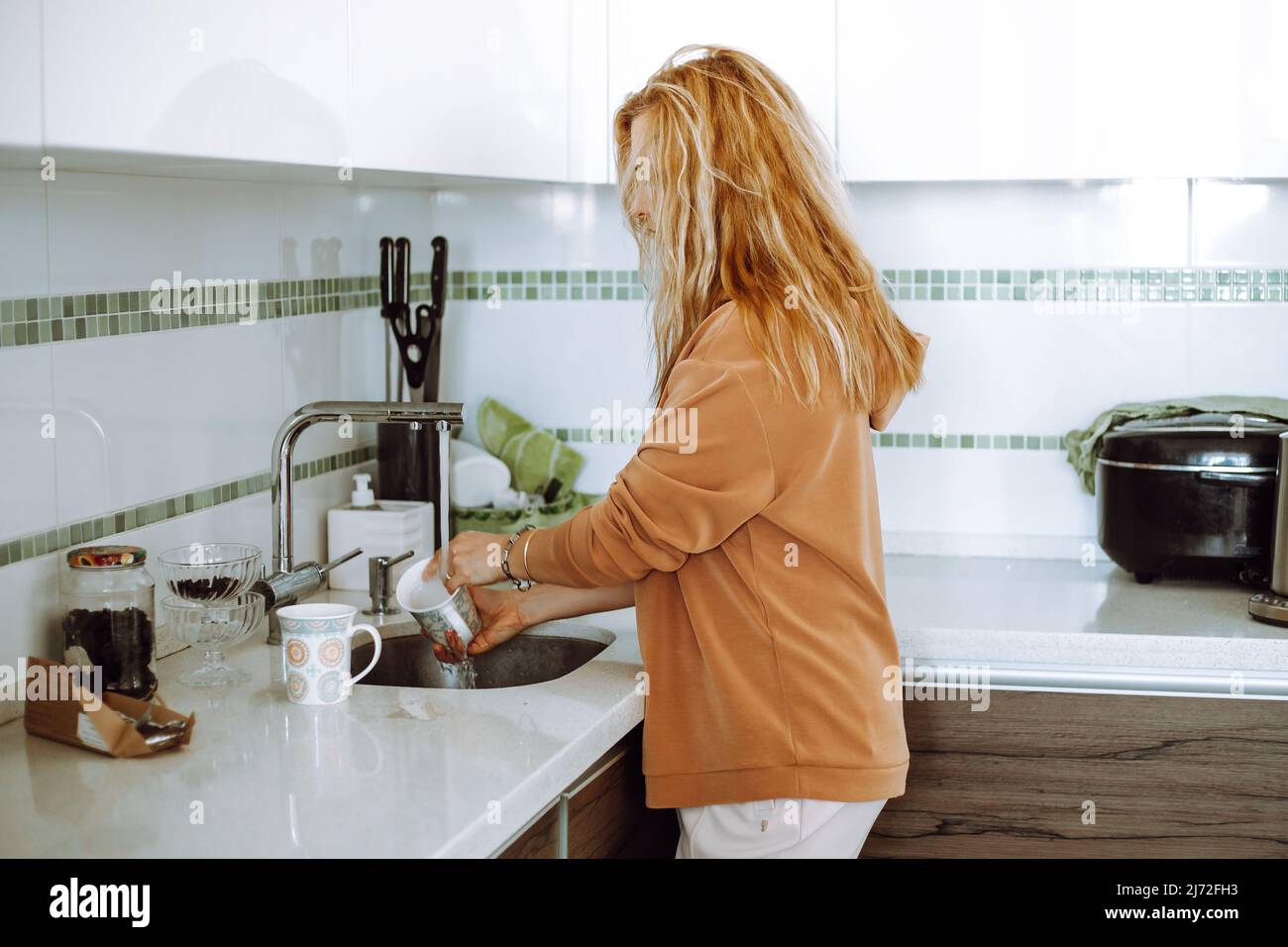 Femme blonde aux cheveux longs dans des vêtements décontractés, laver la vaisselle dans l'évier, dans la cuisine blanche. Femme au foyer et mère à la maison Banque D'Images