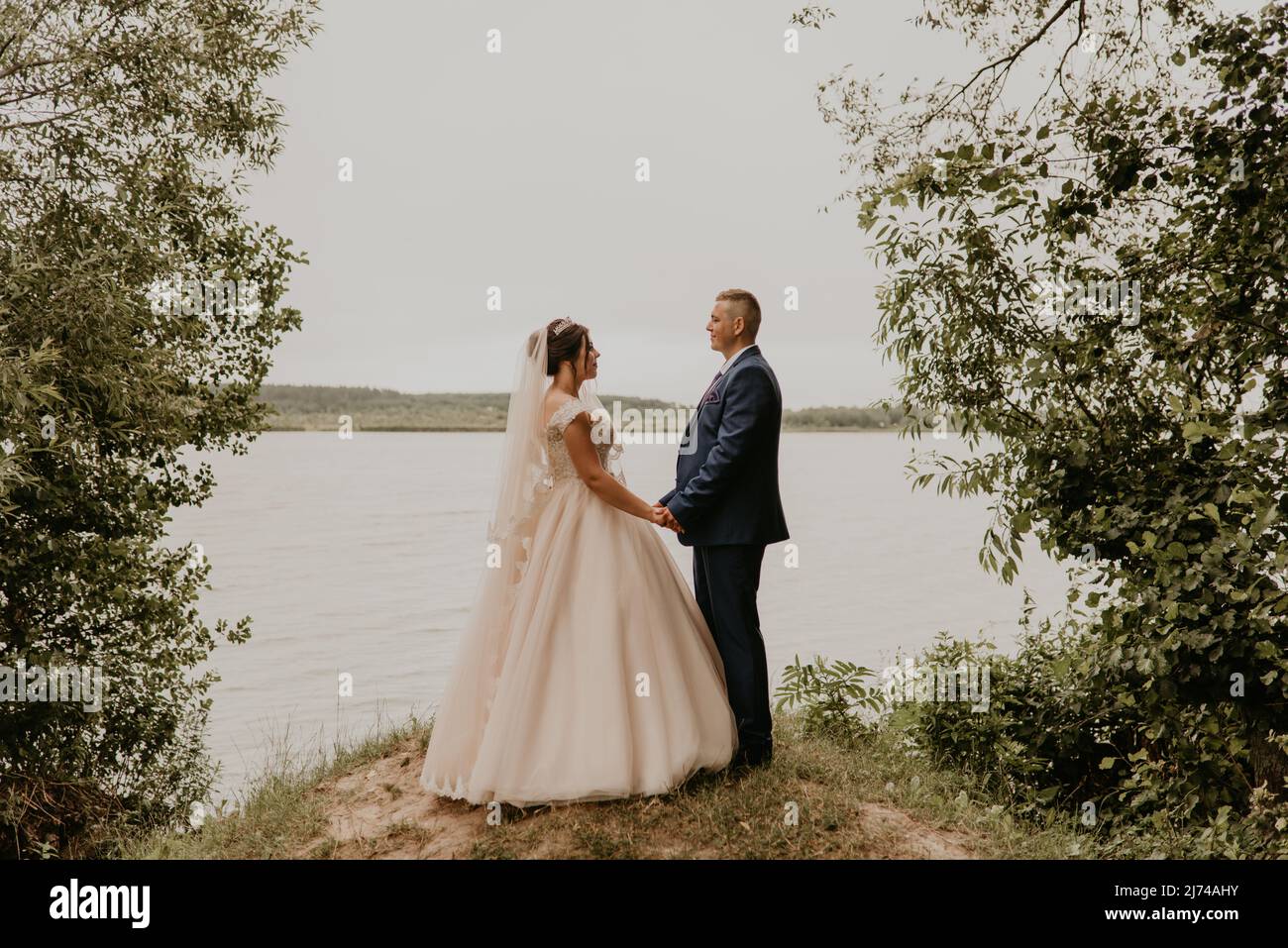 Blonde européenne caucasienne jeune homme marié en costume bleu et femme à cheveux noirs mariée en robe de mariage blanche avec voile long et tiara sur la tête. Nouveaux mariés Banque D'Images