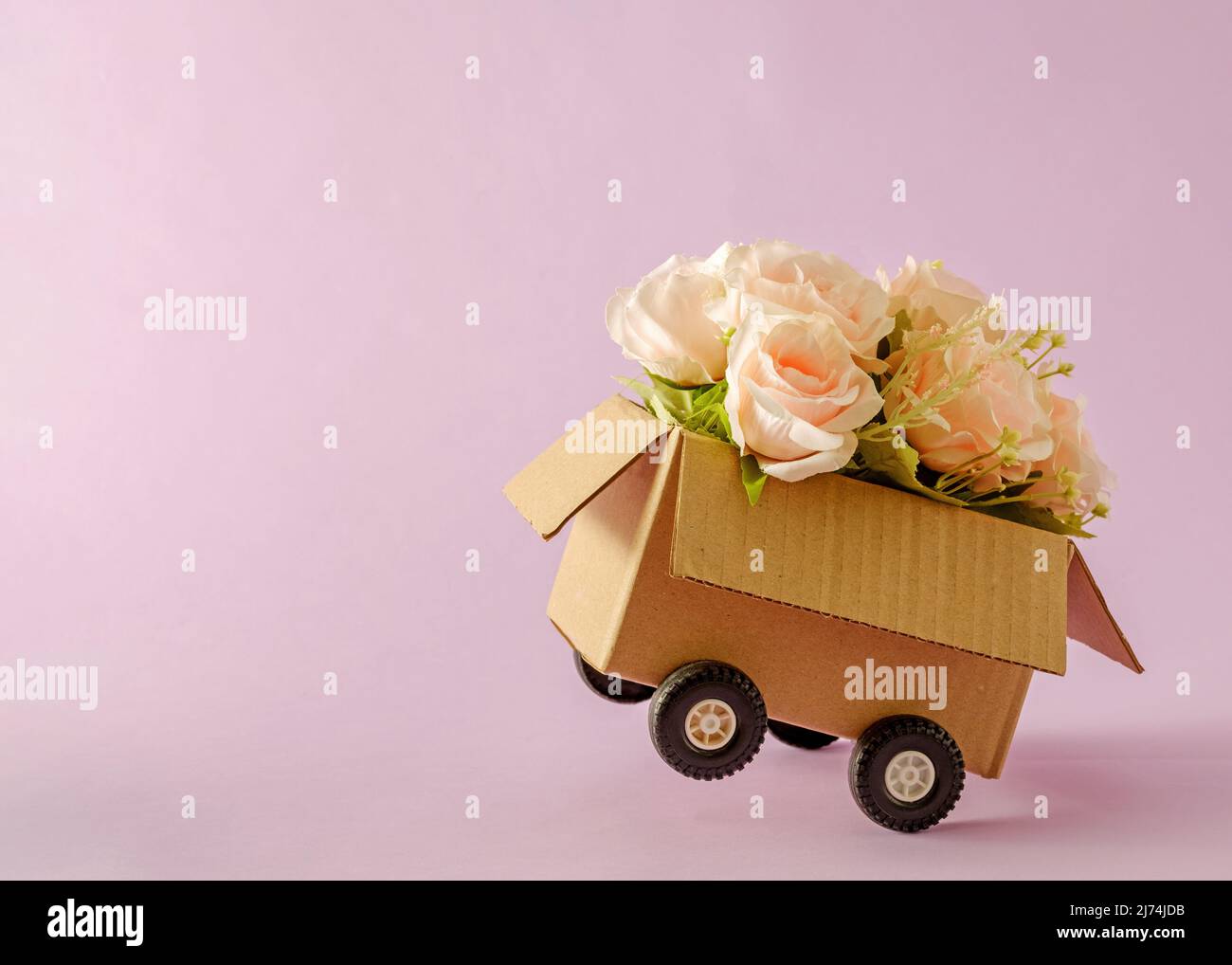 Conteneur de livraison de boîte en carton avec roues de camion et bouquet de roses roses. Concept minimal de courrier et félicitations avec un cadeau de vacances. Banque D'Images