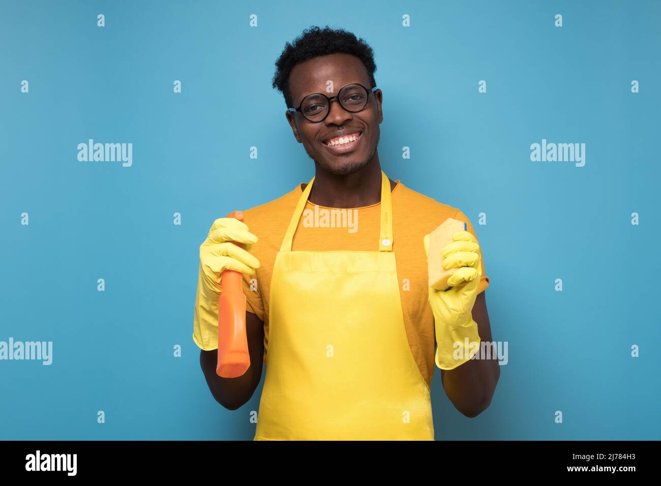 Beau homme africain du service de nettoyage prêt à ranger votre appartement avec un détergent et une éponge. Studio tourné sur un mur bleu Banque D'Images