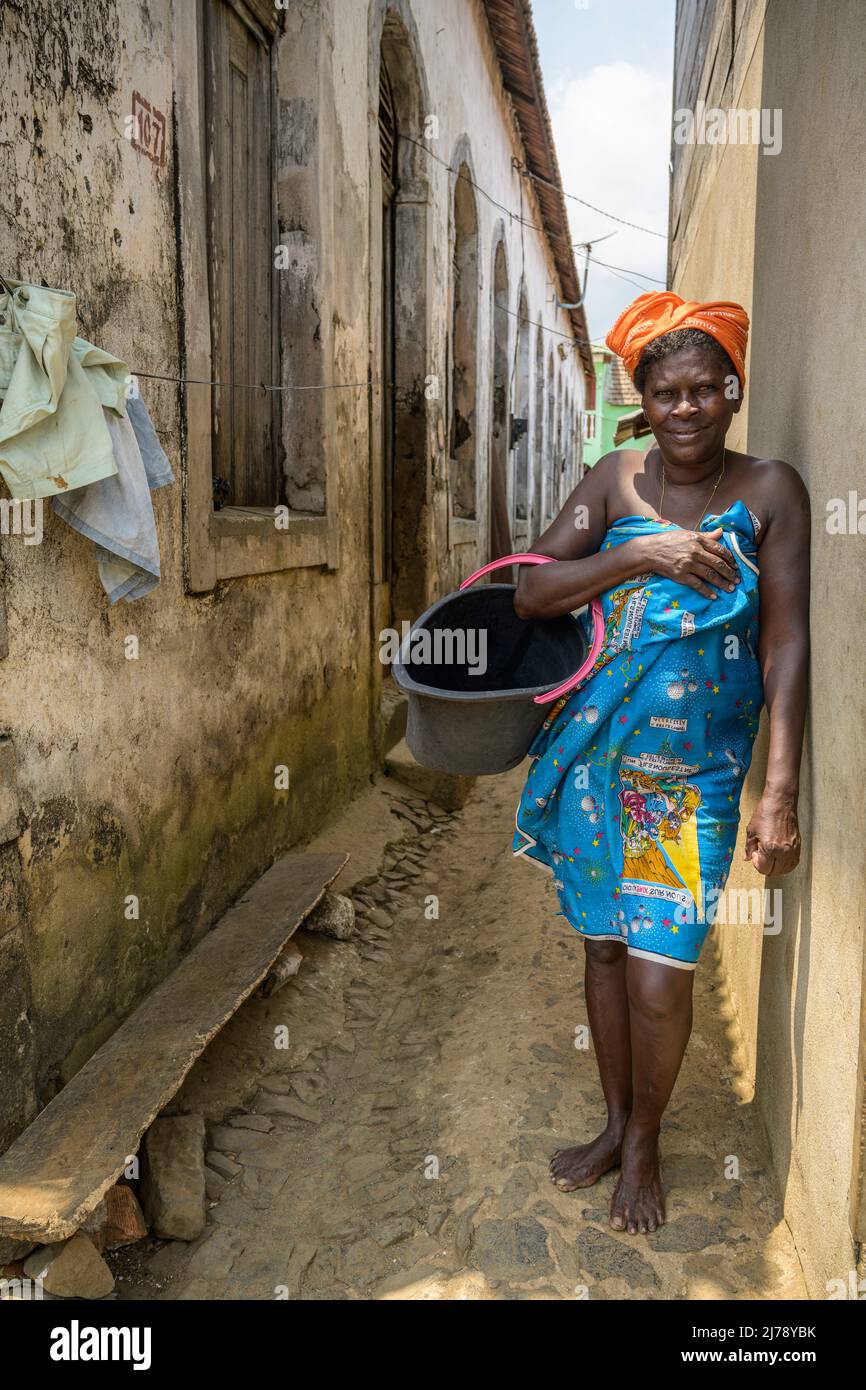 Femme pieds nus avec foulard et pareo avec un seau en plastique dans les rues étroites de l'ancienne plantation coloniale de Roça Agua Ize. Banque D'Images