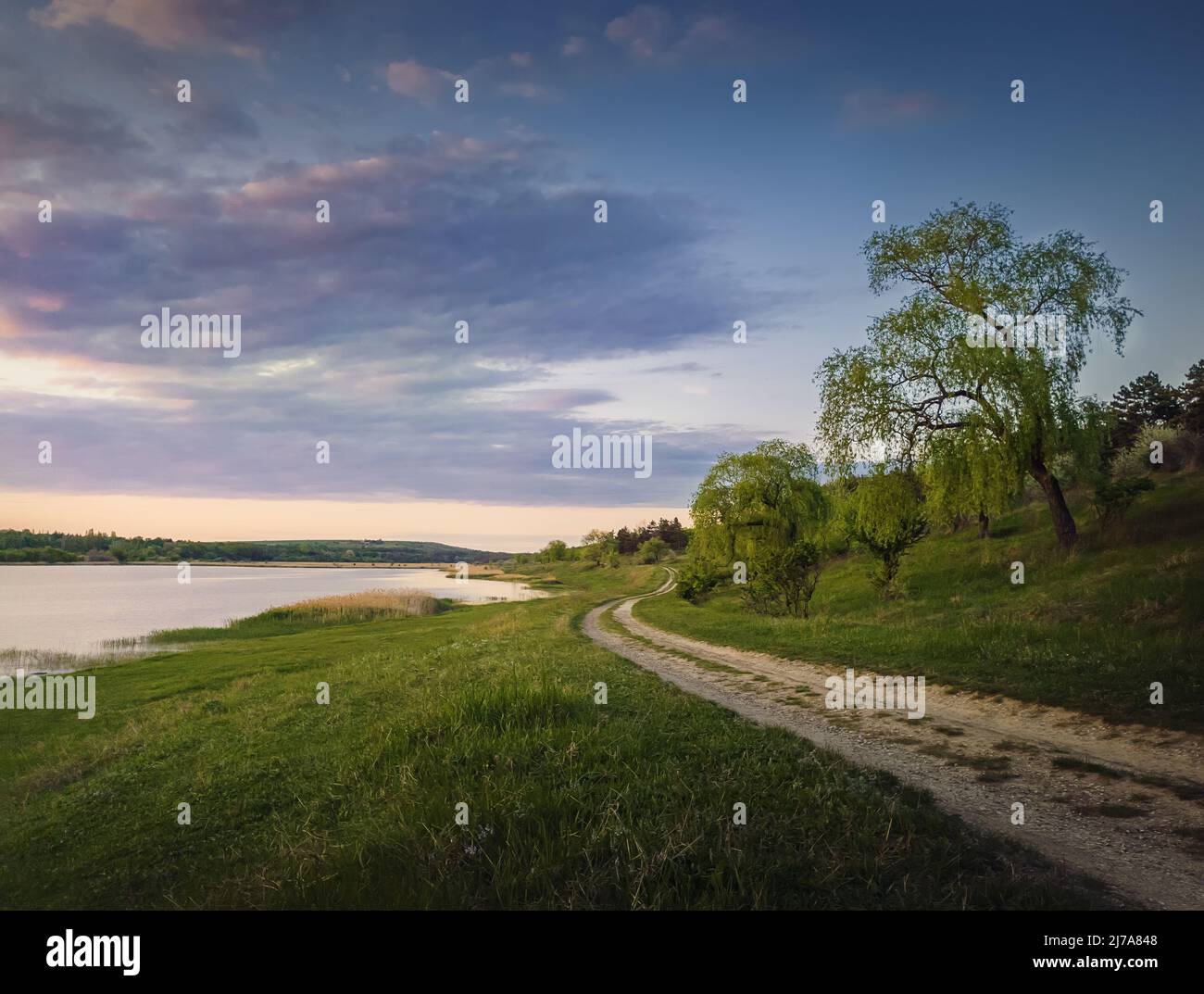 Paysage rural avec une route de campagne entre lac et forêt. Scène estivale idyllique, soirée paisible avec ciel de coucher de soleil coloré Banque D'Images