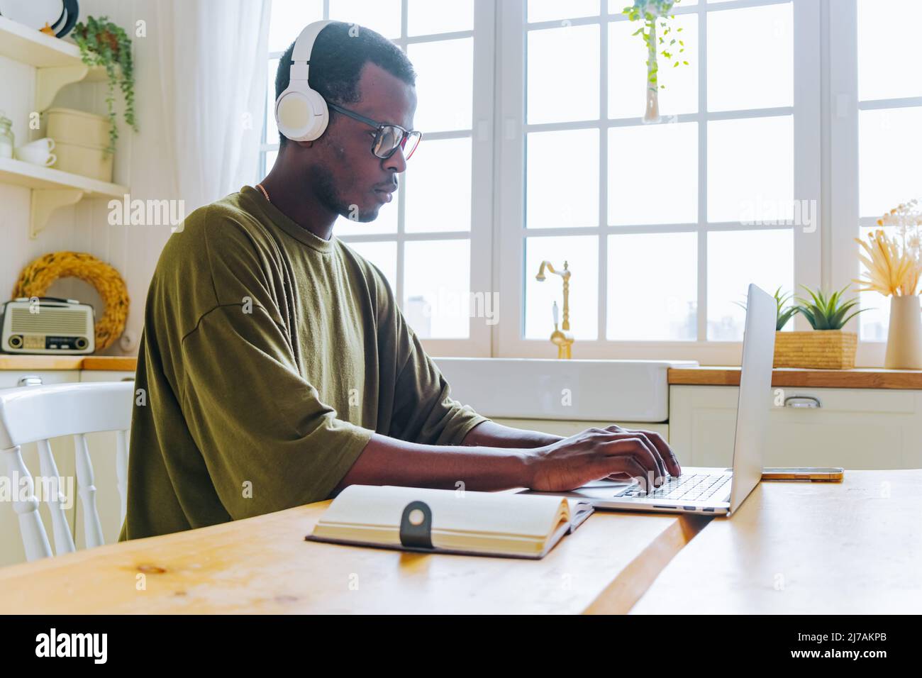 L'homme afro-américain dans le casque prend des cours en ligne via ordinateur portable écrivant des notes. Guy écoute l'audio à la table contre une grande fenêtre Banque D'Images