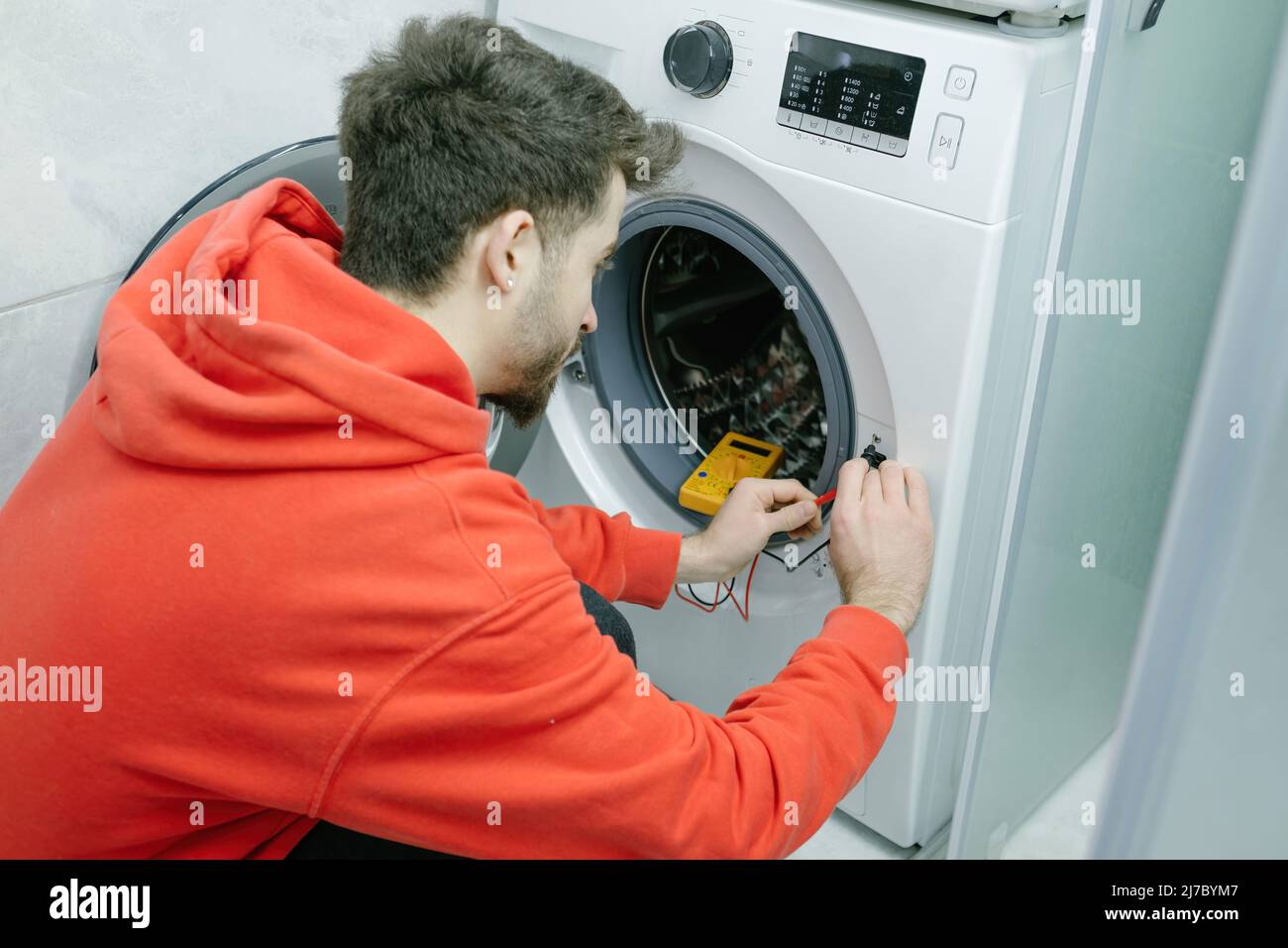 Réparateur, jeune homme beau répare la machine à laver cassée dans sa maison, il vérifie s'il y a de l'électricité avec un voltmètre Banque D'Images