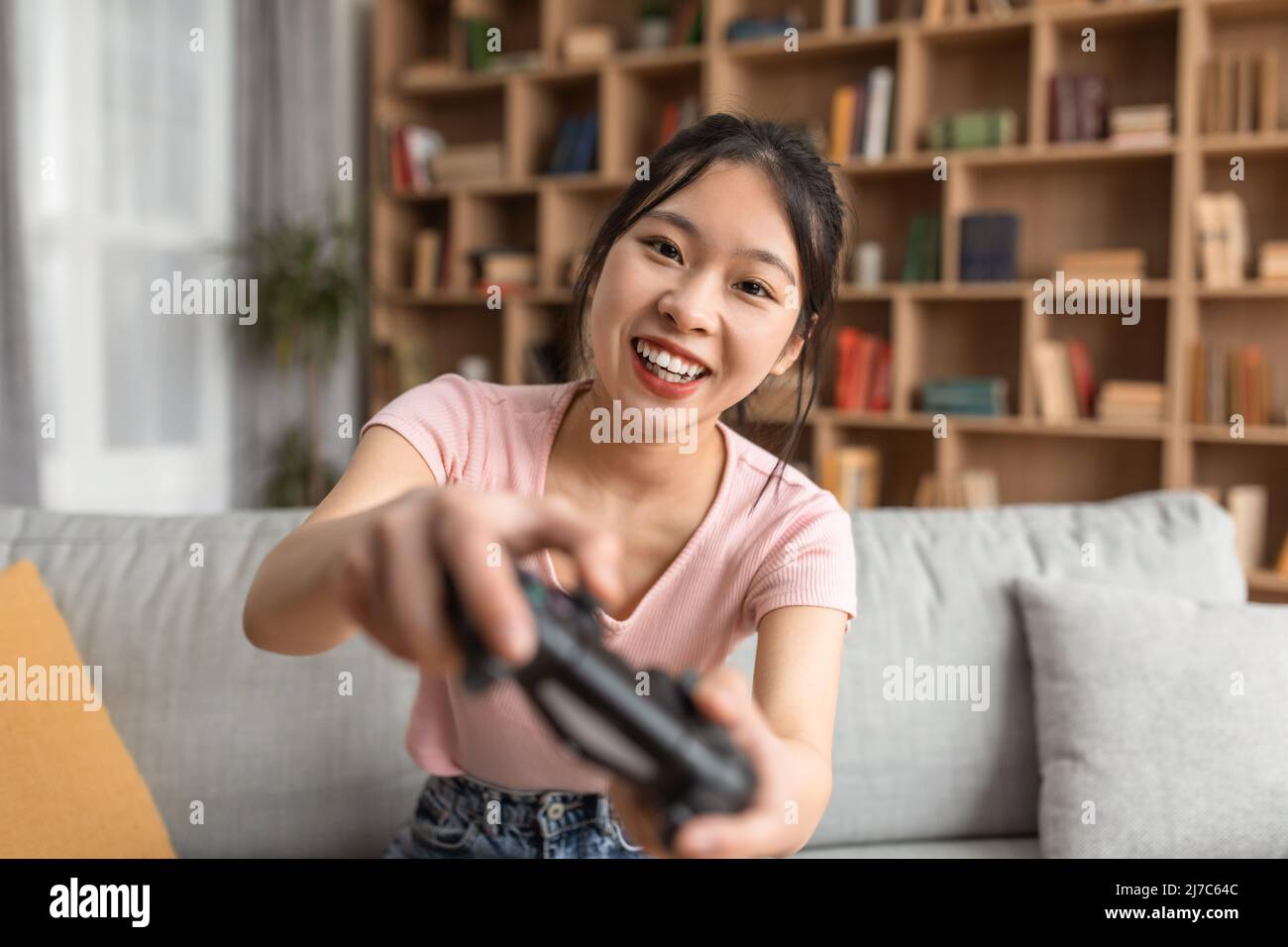 Dame coréenne ludique utilisant le joystick et jouant à des jeux vidéo en ligne, assis sur un canapé dans la salle de séjour, à proximité Banque D'Images