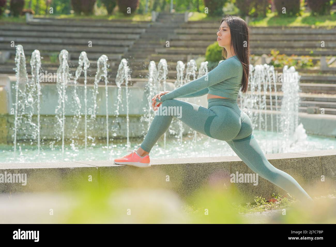 Belle jeune fille de cheveux bruns en vert olive tenue de sport s'étendant après l'exercice de plein air près de la fontaine publique Banque D'Images
