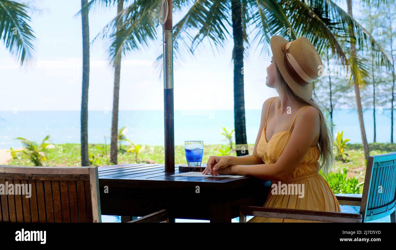 Vacances tropicales. Femme souriante et heureuse dans un chapeau assis dans un café extérieur en bord de mer Banque D'Images