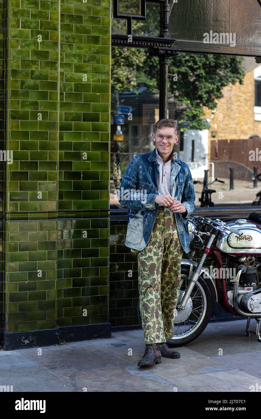 Homme élégant portant un pantalon camouflage sur le côté du pub avec une moto classique Banque D'Images