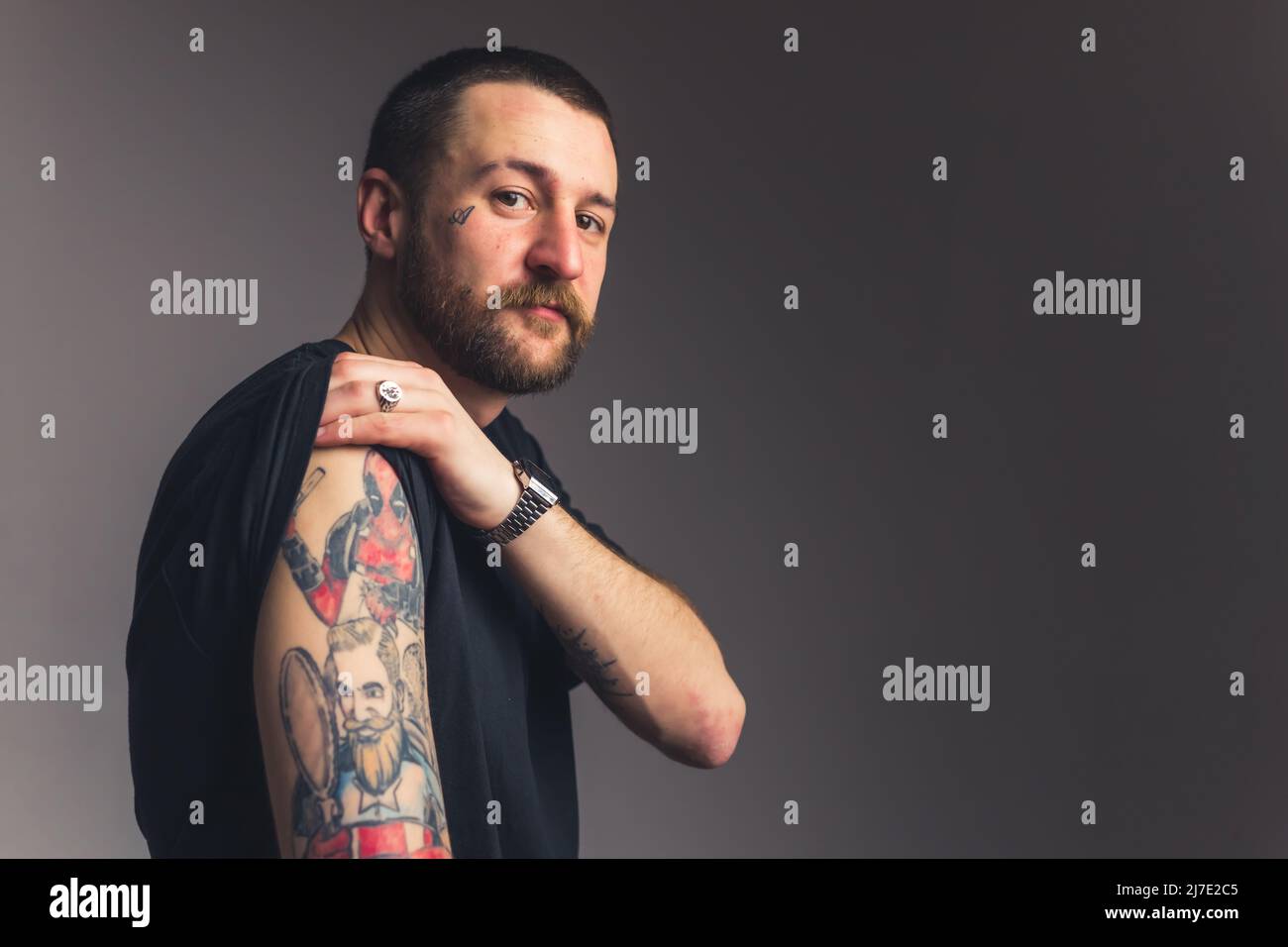 Gangster slouchy homme dans un t-shirt noir montrant à l'appareil photo tatouages sur son épaule sur fond gris foncé. Photo de haute qualité Banque D'Images