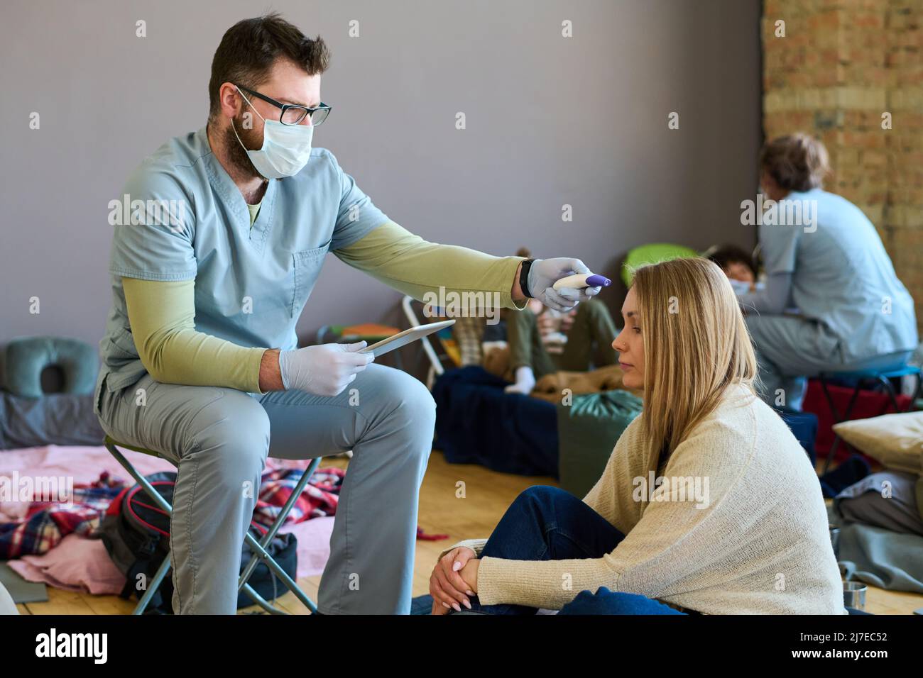 Jeune homme médecin en uniforme, gants et masque de protection mesurant la température de la femme blonde réfugiée vivant temporairement dans le camp Banque D'Images